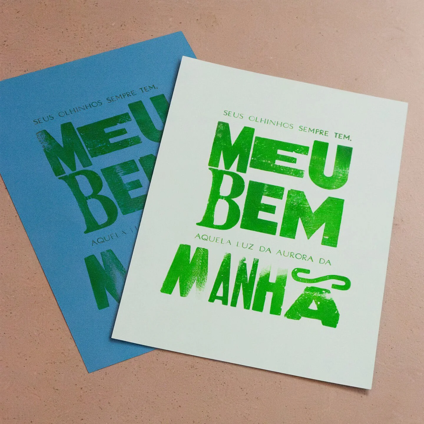 Two printed sheets of paper with green text, one on a blue background and the other on a white background, containing the phrase 'SEUS OLHINHOS SEMPRE TEM. MEU BEM AQUELA LUZ DA AURORA DA MANHÃS'.