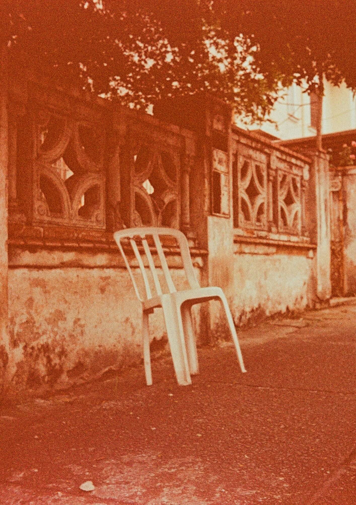 A white plastic chair leaning against a weathered brick wall with ornate windows, a tree with full foliage in the background, and a chair on an unpaved dirt ground, all bathed in a reddish-orange hue.