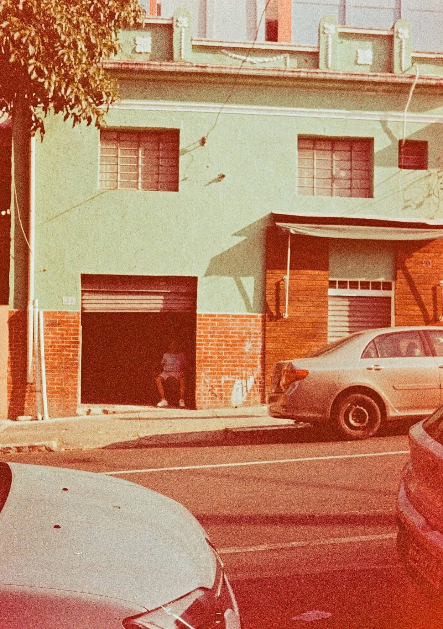A street scene showing parked cars in front of a building with a green upper facade and a brown brick lower facade. There is a person in a pink hat and backpack standing near an open garage door.