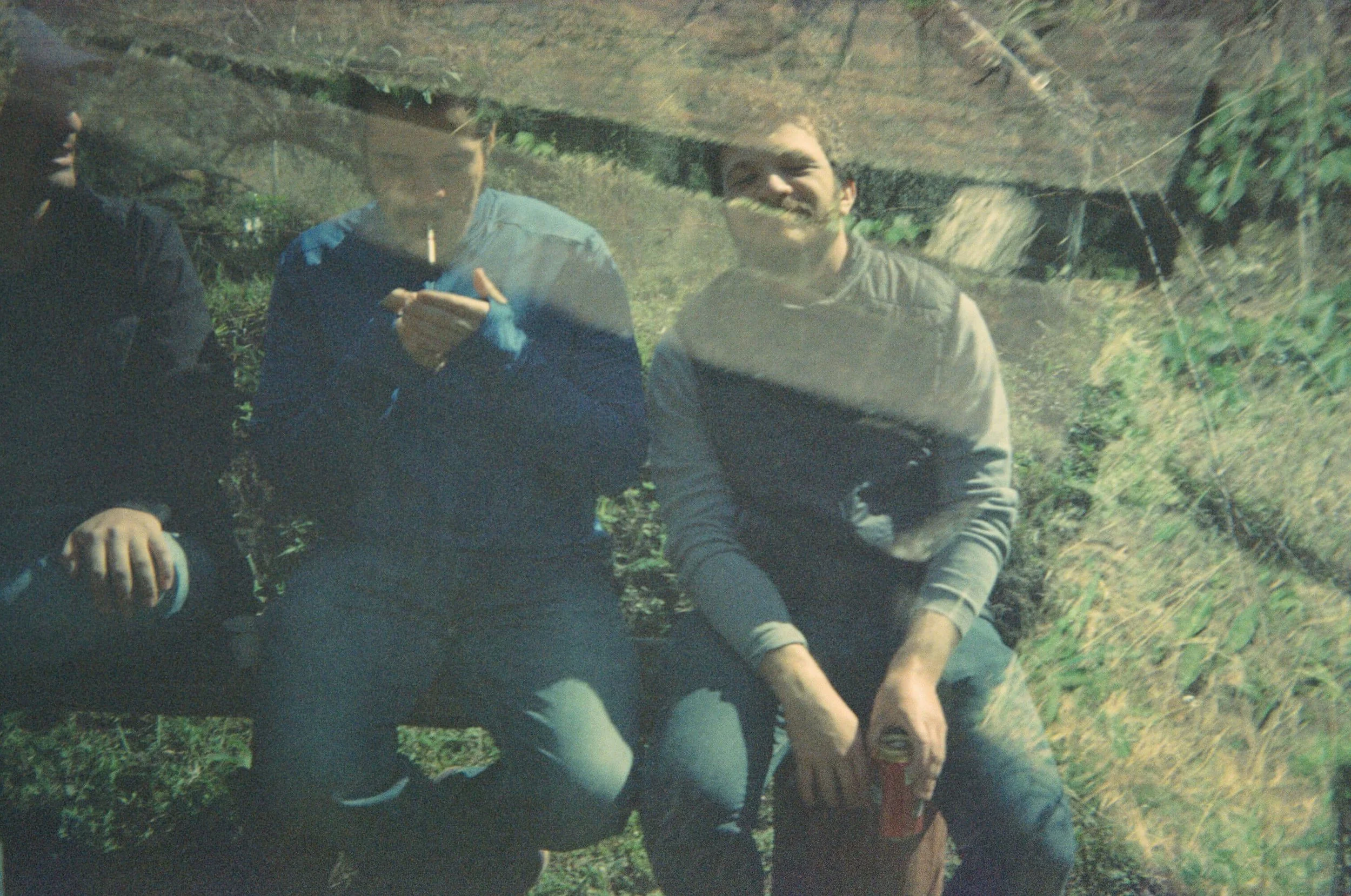 Three people are outdoors, with two of them visible and sitting or lying on the grass. One person is wearing a gray and black striped hoodie, and another is wearing a blue jacket. The image has a reflection or distortion that makes it appear as if th