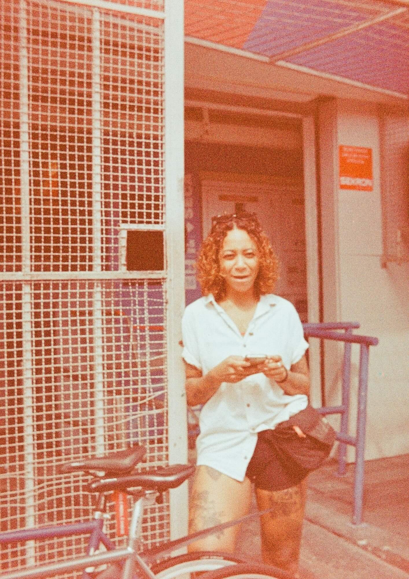 A woman with curly hair sitting at an airport gate, looking at her phone, surrounded by metal structures and seating areas.