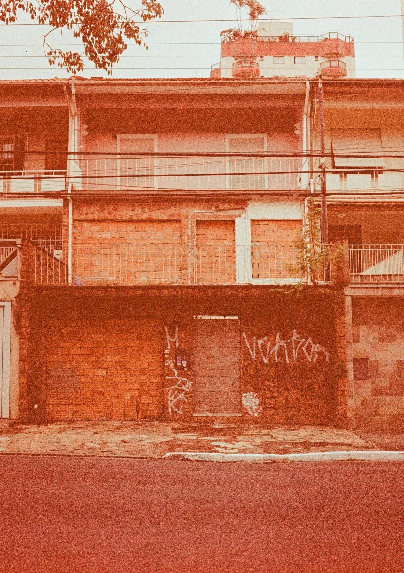 A three-story building with a brick facade on the ground floor and beige walls on the upper floors. The building has metal railings on the balconies and windows with closed shutters. There is graffiti on the garage door and some plants growing along 