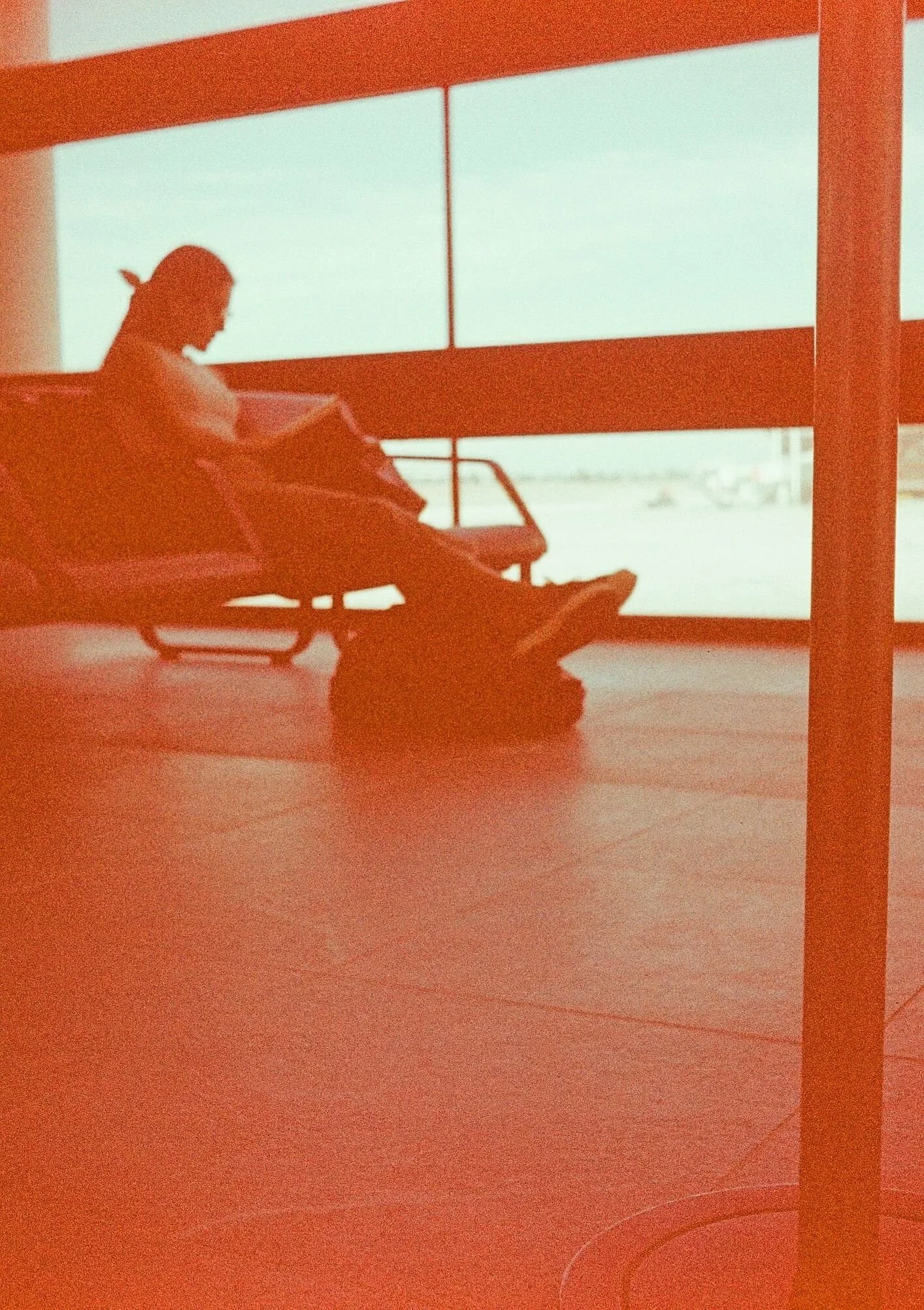 Silhouette of a person sitting on a bench, reading a book, inside a room with large windows and a view of an airport tarmac outside.