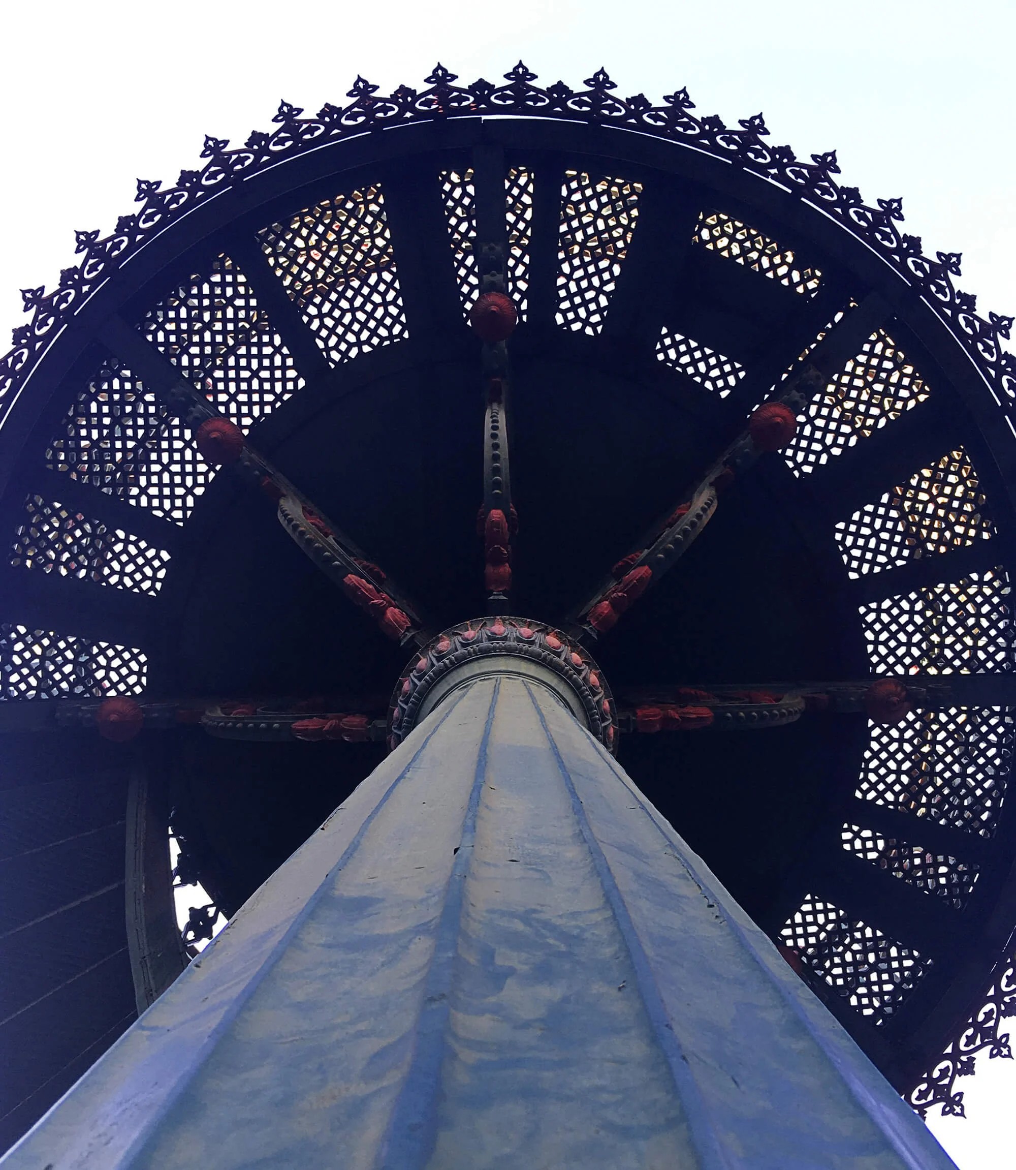 The photo shows an upward view of a decorative Ferris wheel structure with intricate lattice and ornate patterns, lit with small lights against a pale sky.