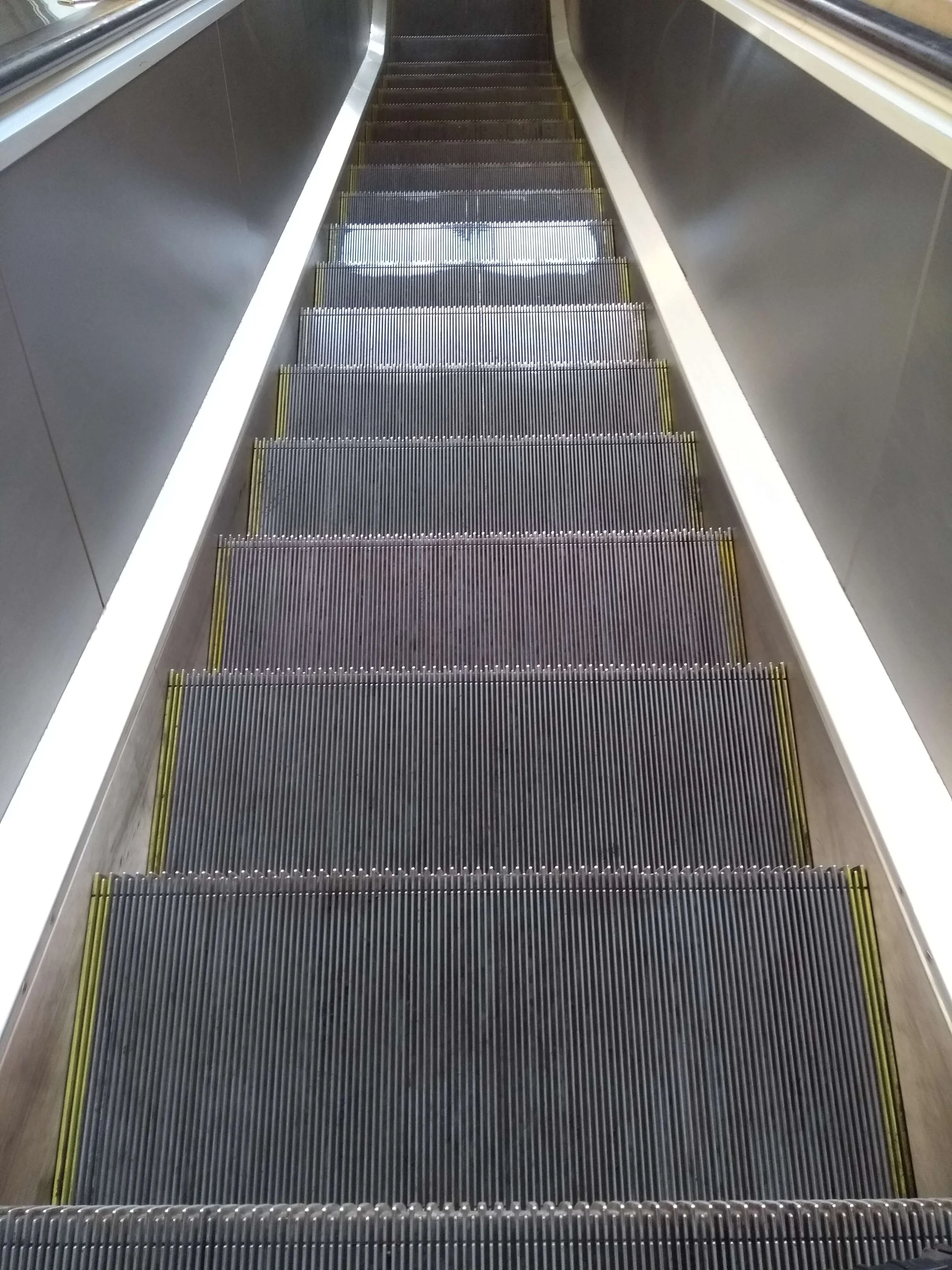Looking down an escalator in a modern building, metal steps with yellow safety lines on the sides, flanked by sleek, dark-colored handrails and walls.