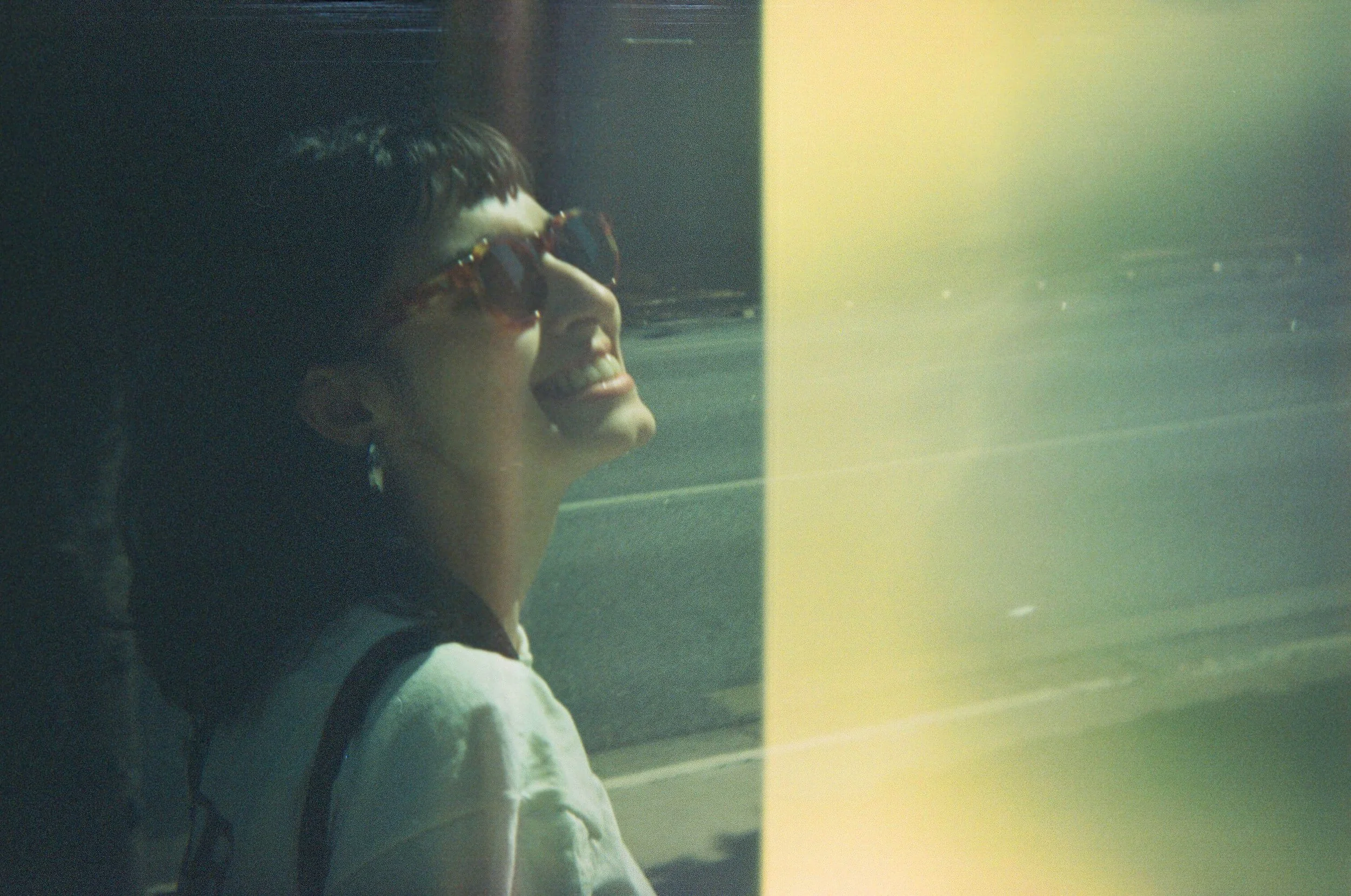 A person wearing sunglasses smiling, partially visible, with their face turned slightly to the right, sitting near a window with sunlight.