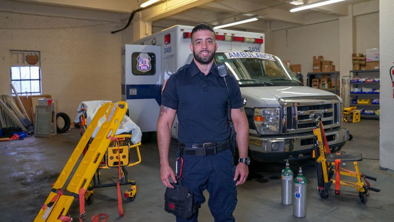 A male EMS worker standing inside a garage with an ambulance and medical equipment around him, including a stretcher and oxygen tanks.