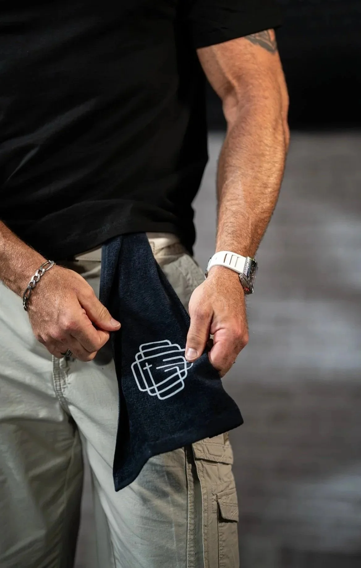 A person wearing a black shirt, beige cargo shorts, holding a black gym towel with a white logo on it, in an indoor setting.