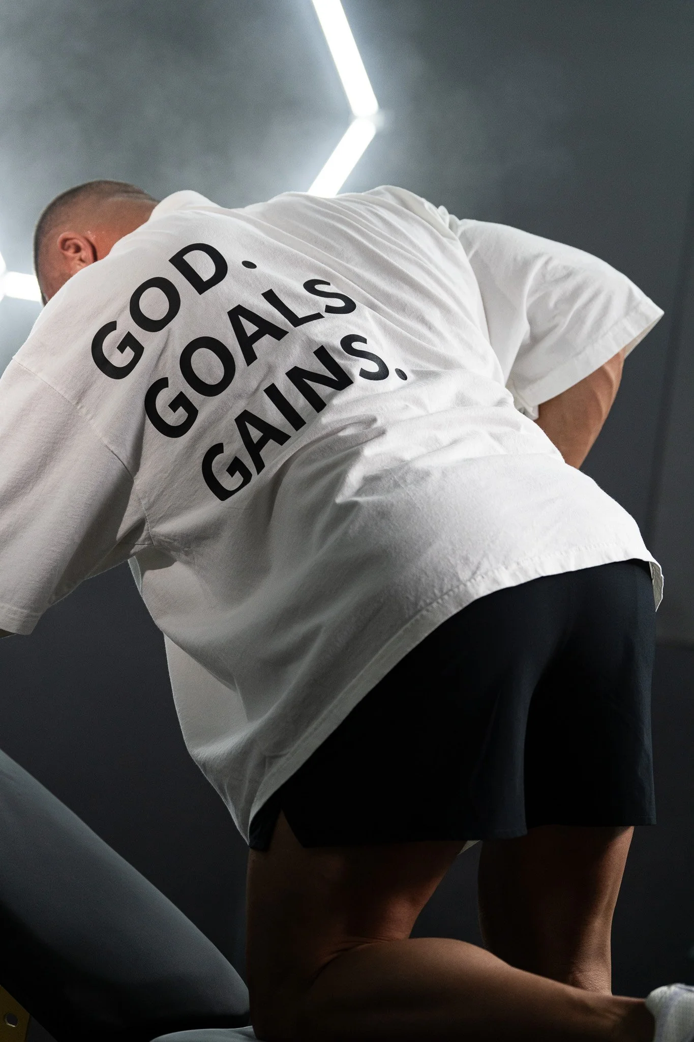 A man is bending over, doing single-arm dumbbell rows, with a white t-shirt that has the words 'GOD. GOALS. GAINS.' written on the back, in a room with dark walls and fluorescent lighting.