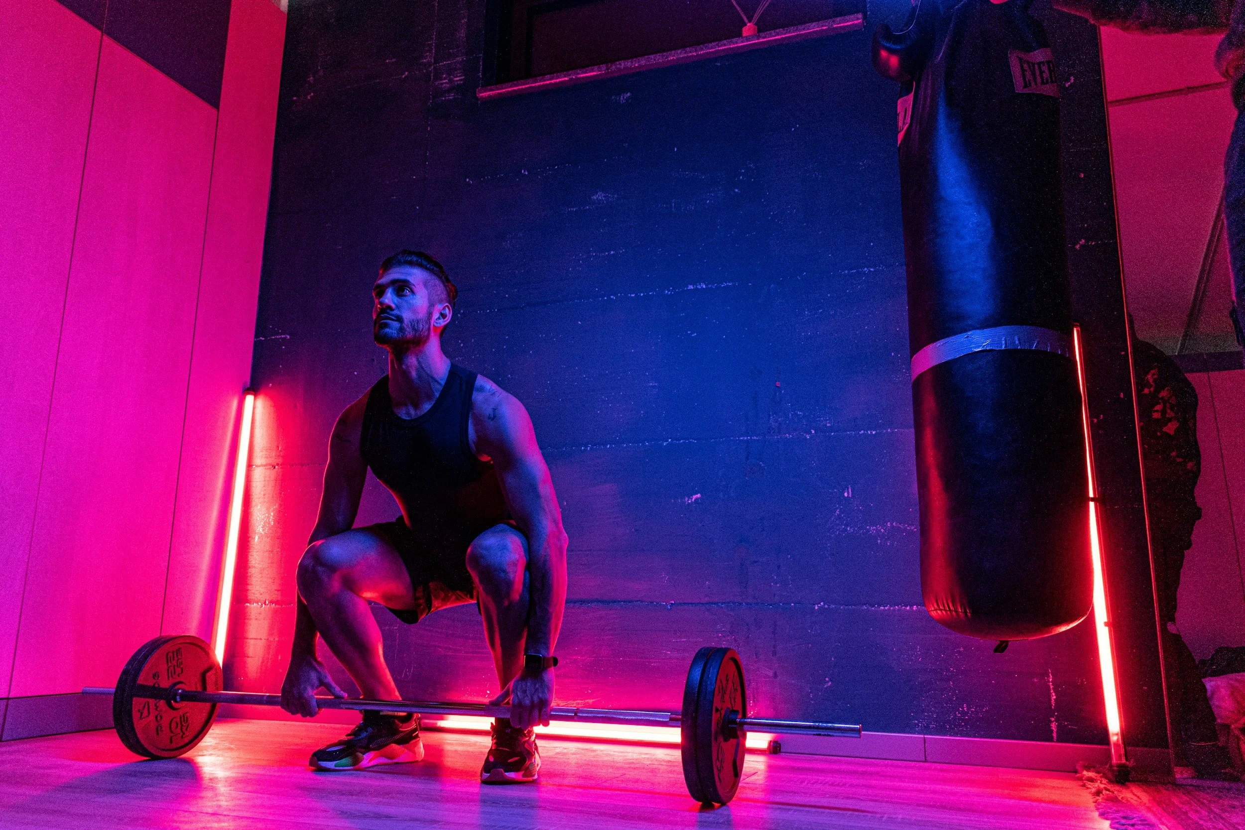 A man in athletic wear doing a deadlift exercise with a barbell in a dimly lit gym with pink and blue neon lighting.