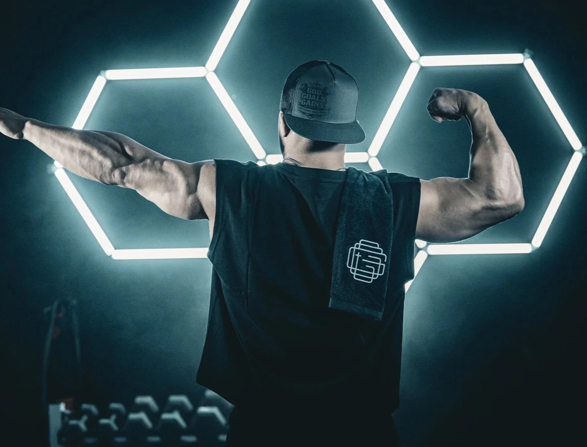Man flexing muscles in front of illuminated hexagon-shaped light installation, wearing a baseball cap and sleeveless shirt.