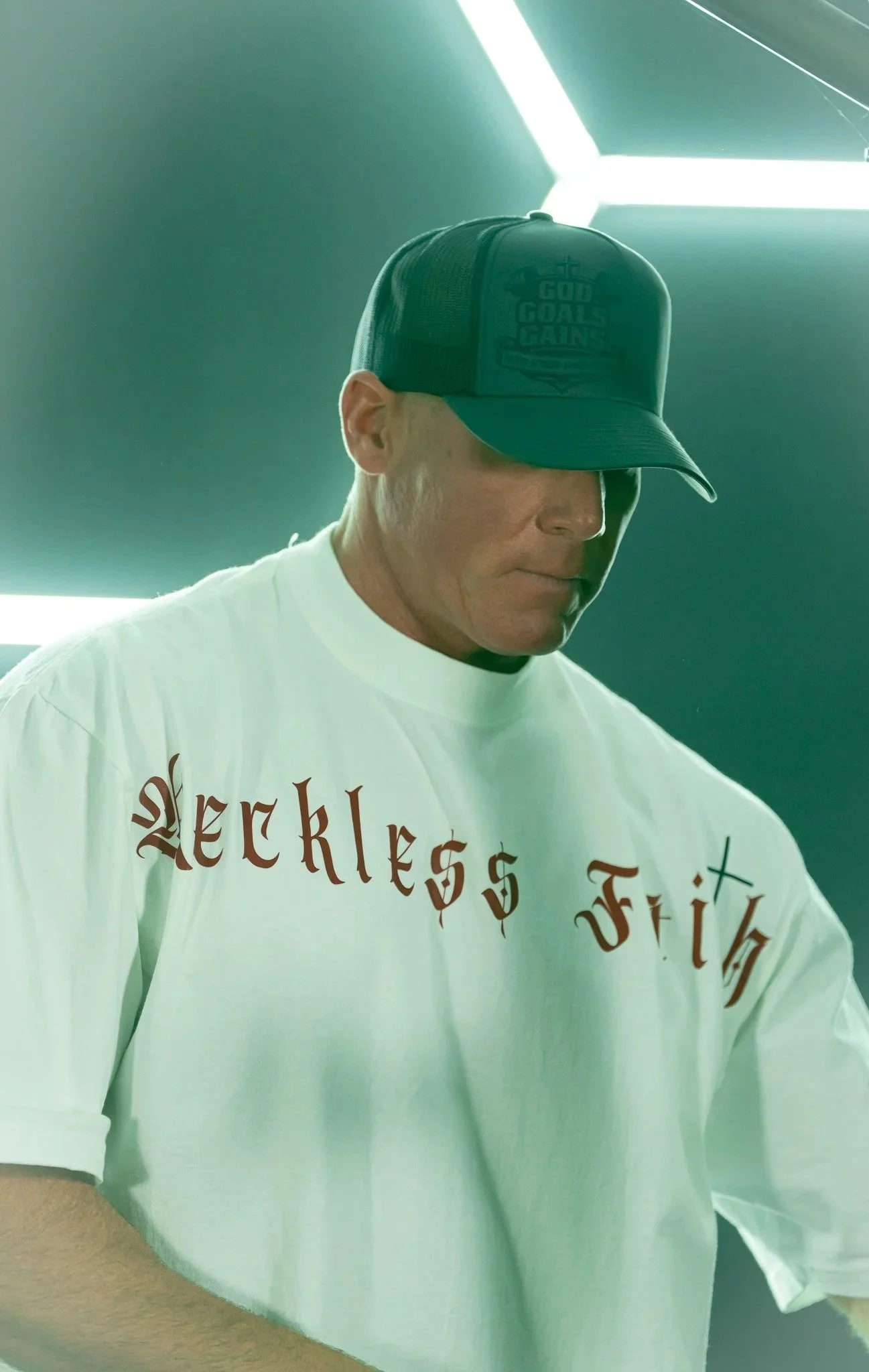A person wearing a dark baseball cap with the words 'God Goals Gains' embroidered on it, and a white T-shirt with a partial phrase in a medieval-style font visible.
