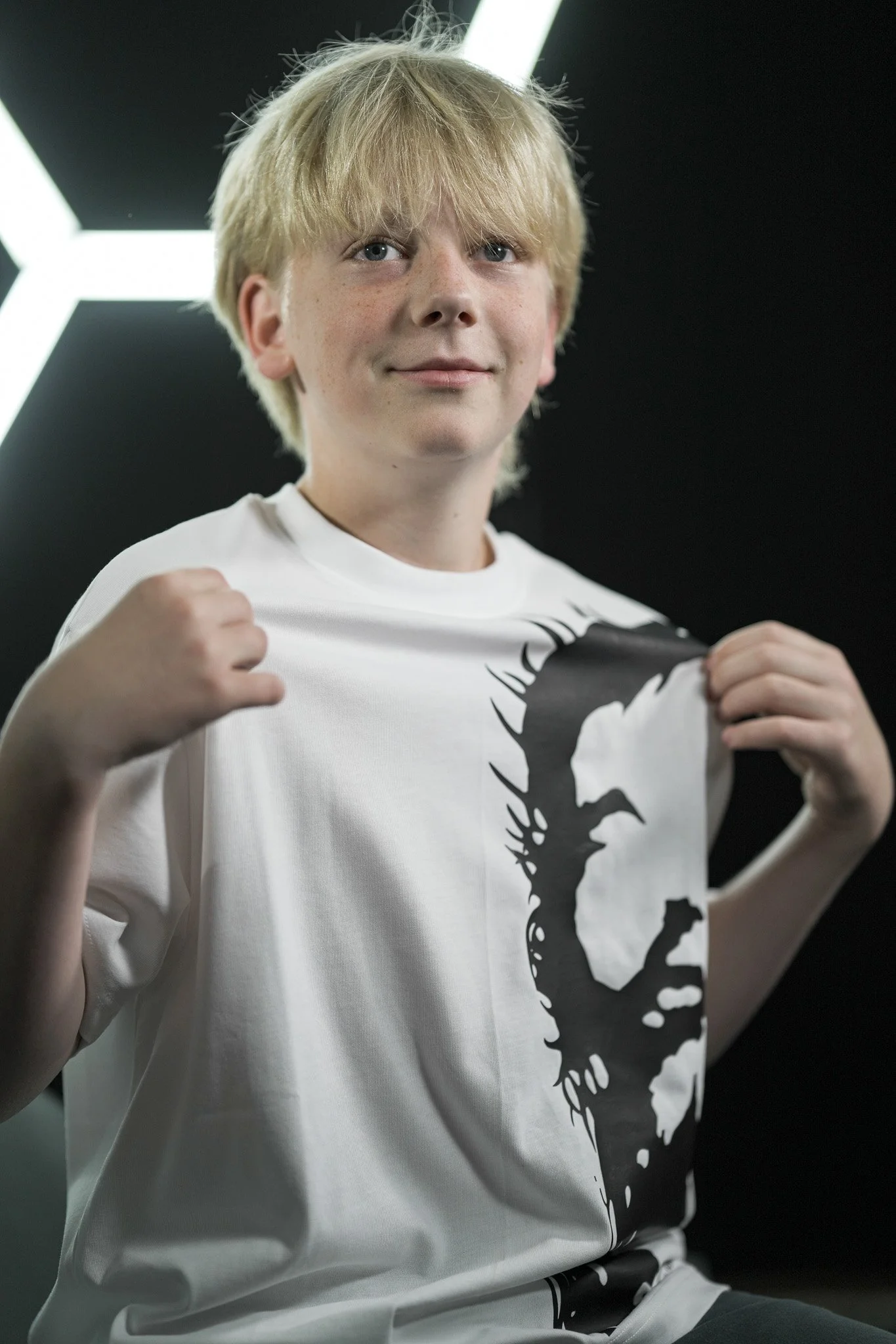 A young person with blonde hair wearing a white T-shirt with a black graphic design, posing with fists raised in a boxing stance, indoors with a dark background and bright overhead lighting.