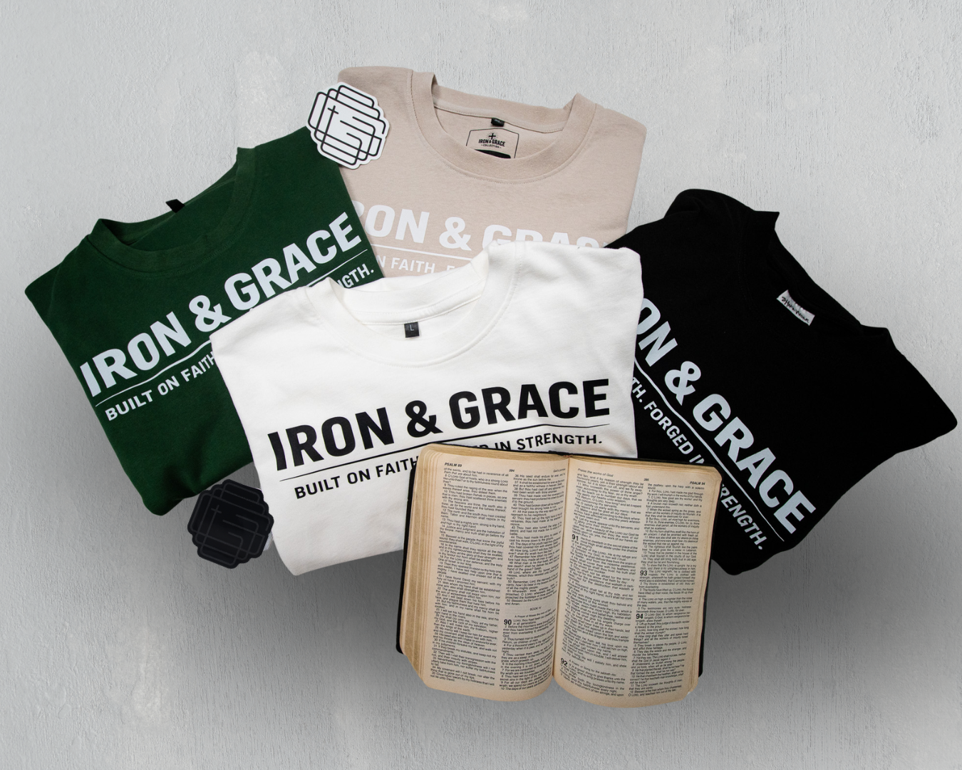 Four folded t-shirts in green, white, beige, and black with the text 'IRON & GRACE' and 'BUILT ON FAITH AND STRENGTH' laid out on a flat surface, with an open Bible in front of the white t-shirt.