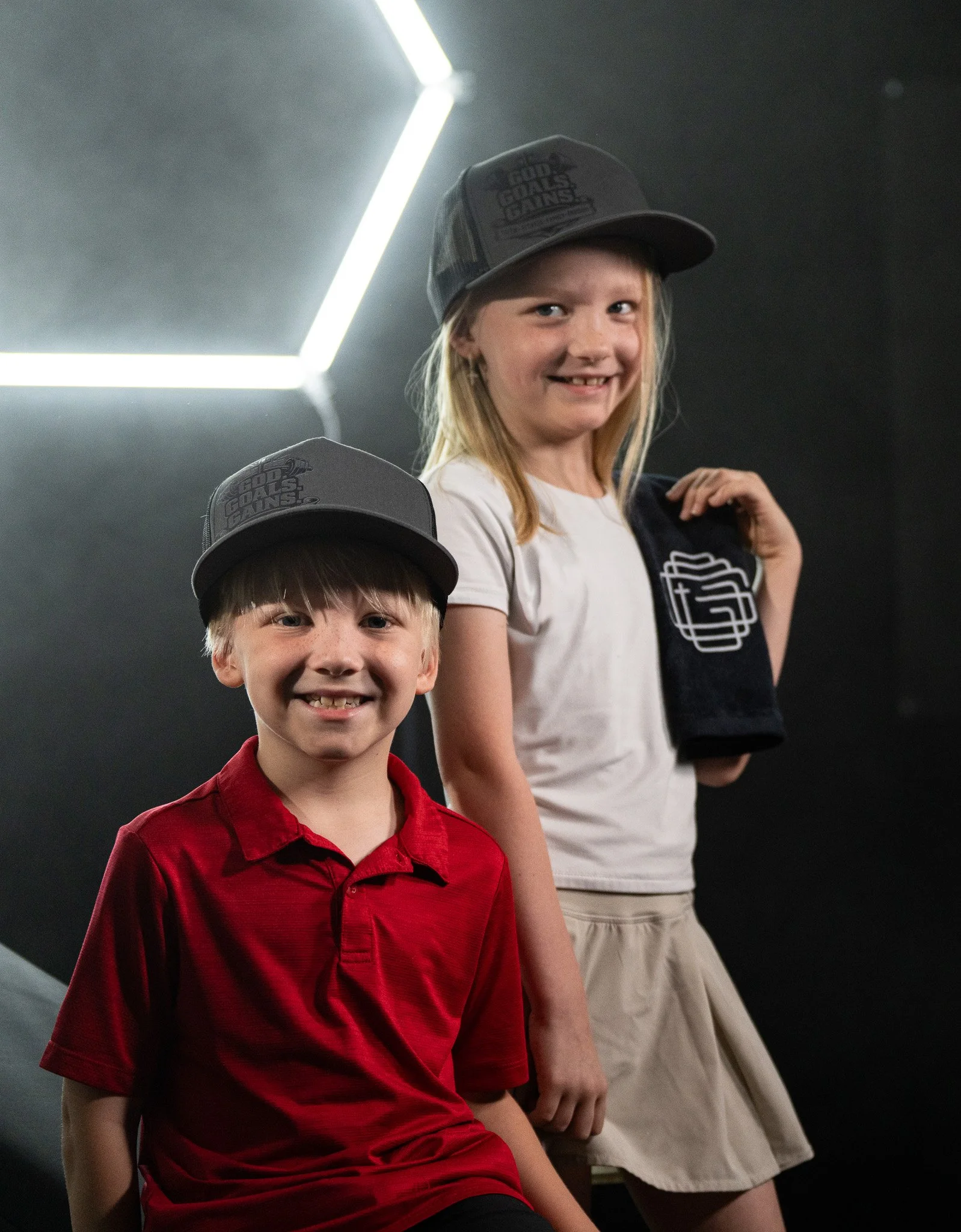 Two children, a boy in a red shirt and a girl in a white shirt, wearing grey hats with the words 'God Causes Giants' printed on them. They are standing in a dark indoor setting, smiling at the camera.