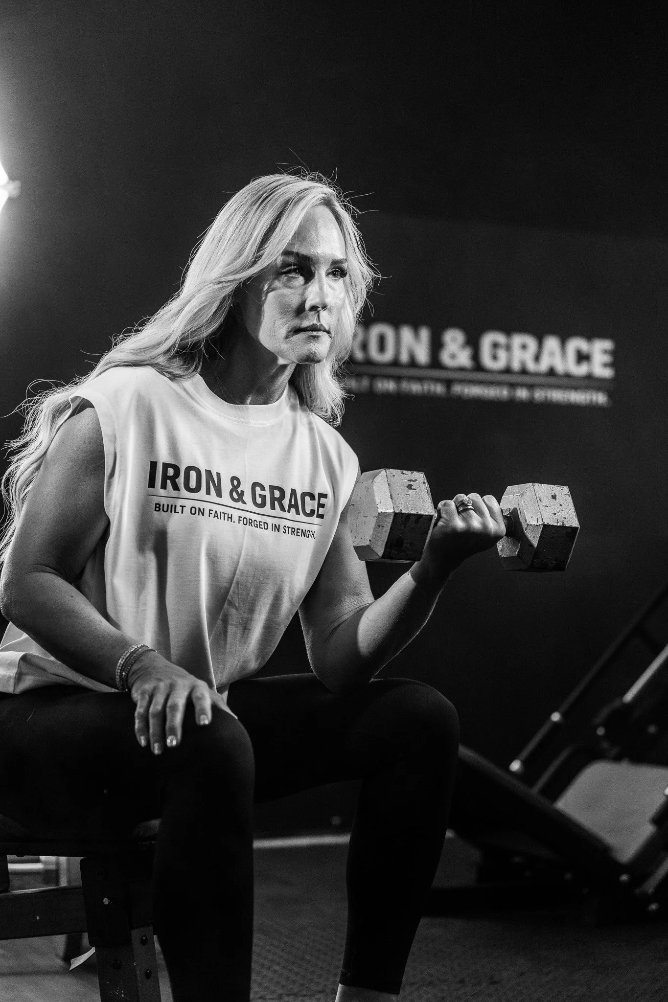 A woman with long blonde hair lifting a dumbbell during a workout at a gym, wearing a sleeveless T-shirt with 'IRON & GRACE' printed on it.
