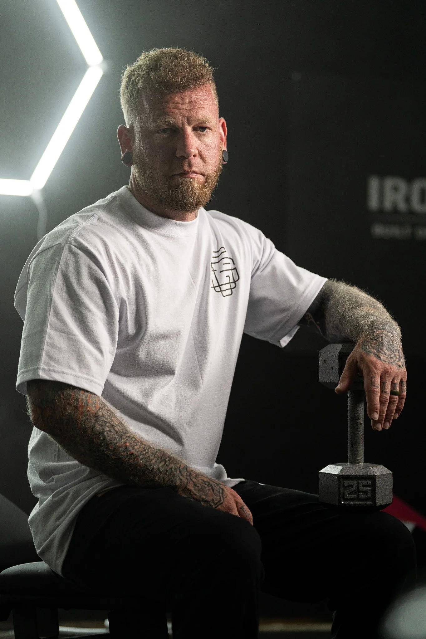 A man with tattoos on his arms sitting in a gym, holding a dumbbell, with a dark background and bright lighting on his face and upper body.