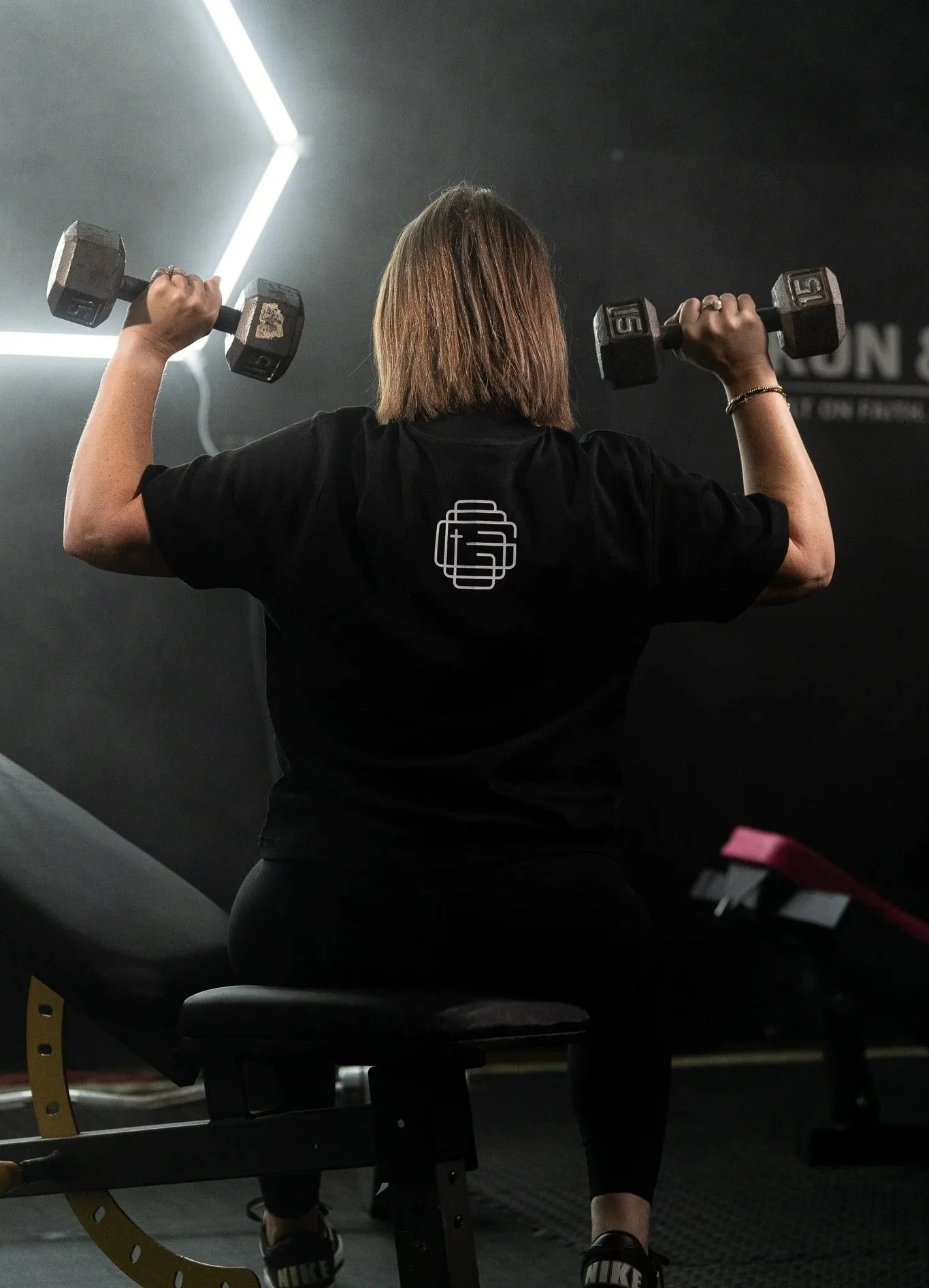 Back of a woman with shoulder-length hair lifting dumbbells in a gym. She is wearing a black shirt with a logo on the back and black sneakers. She is seated on a workout bench.