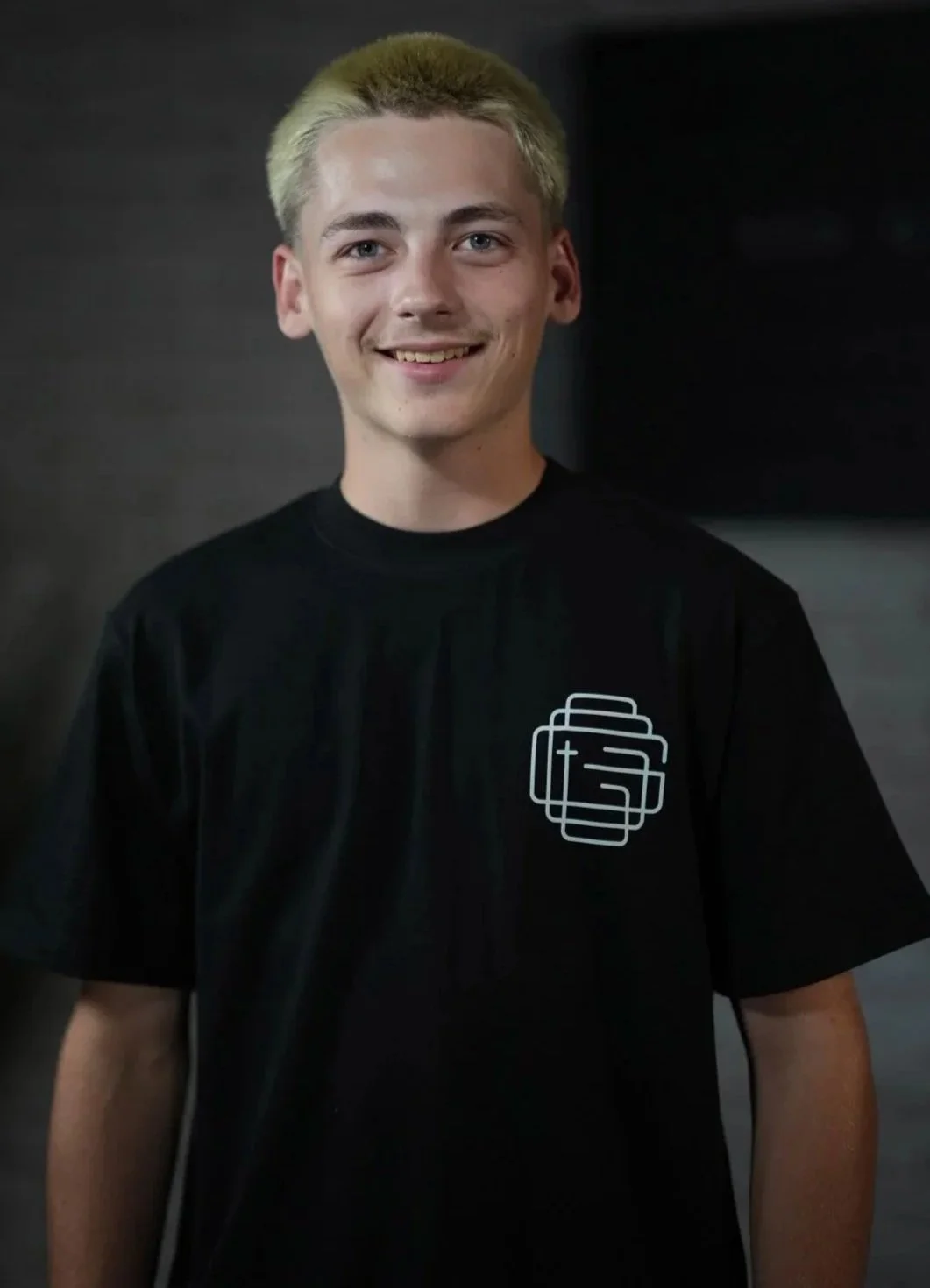 Young man with dyed blonde hair smiling, wearing a black T-shirt with a geometric logo on the left side.