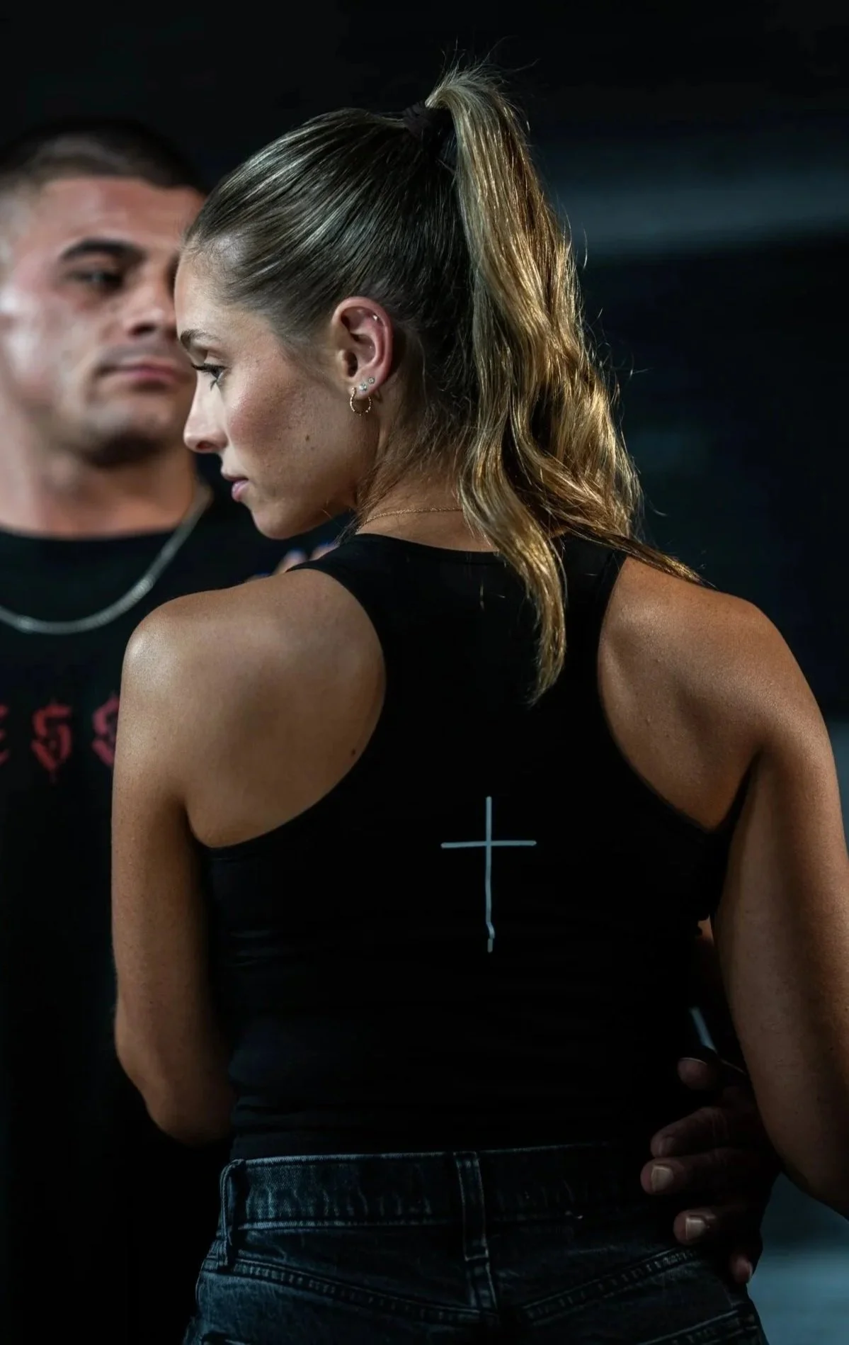 A woman with wavy light brown hair tied in a high ponytail, wearing a black tank top with a white cross on the back, standing next to a man in a black shirt. The woman is in profile, looking downward, with the man partially visible in the background.