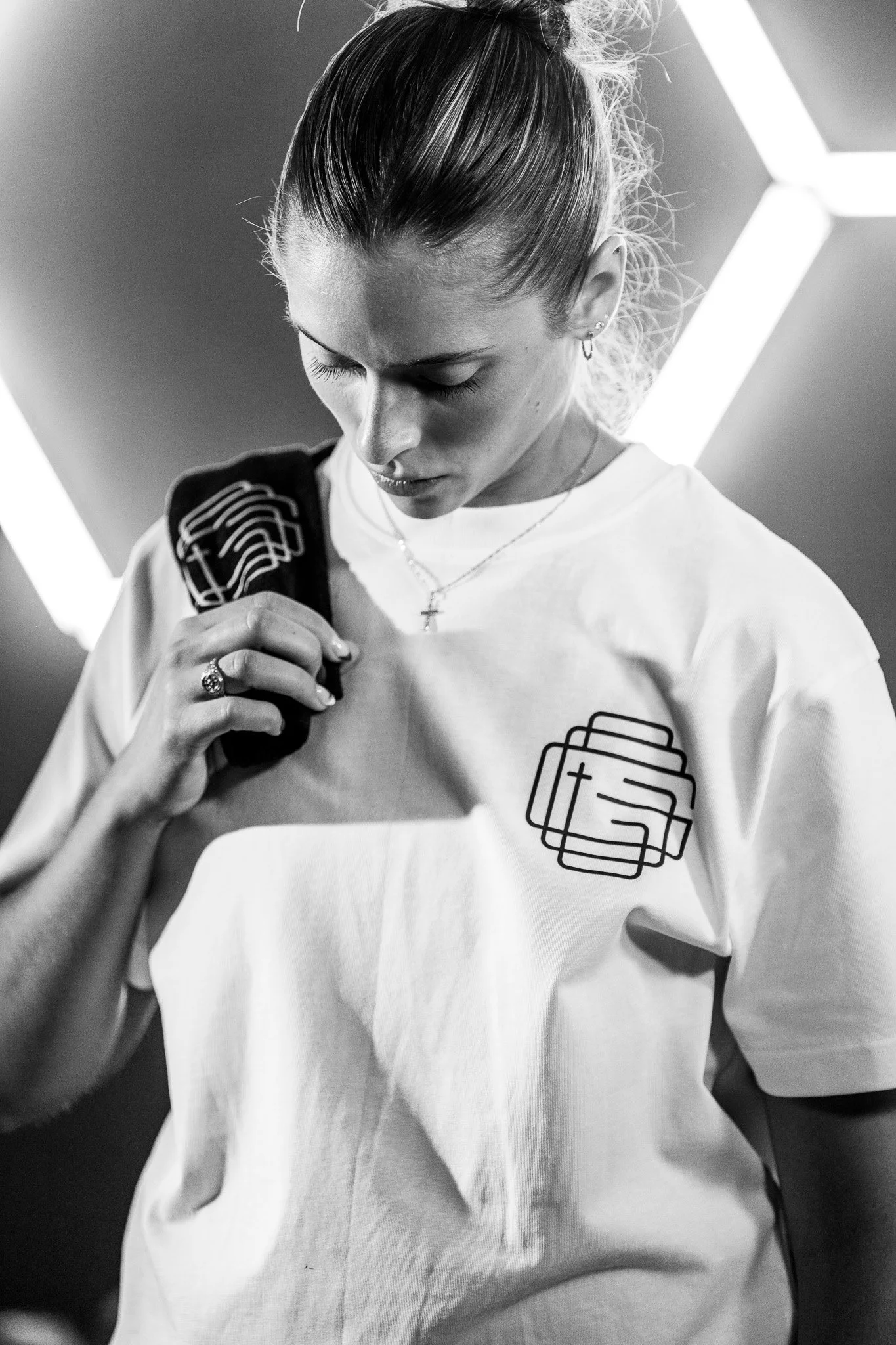 Black and white photo of a woman looking down while holding a black gym towel on her shoulder, wearing a white T-shirt with a graphic design, and a cross necklace. She has earrings and her hair is tied back. The background shows geometric lights.