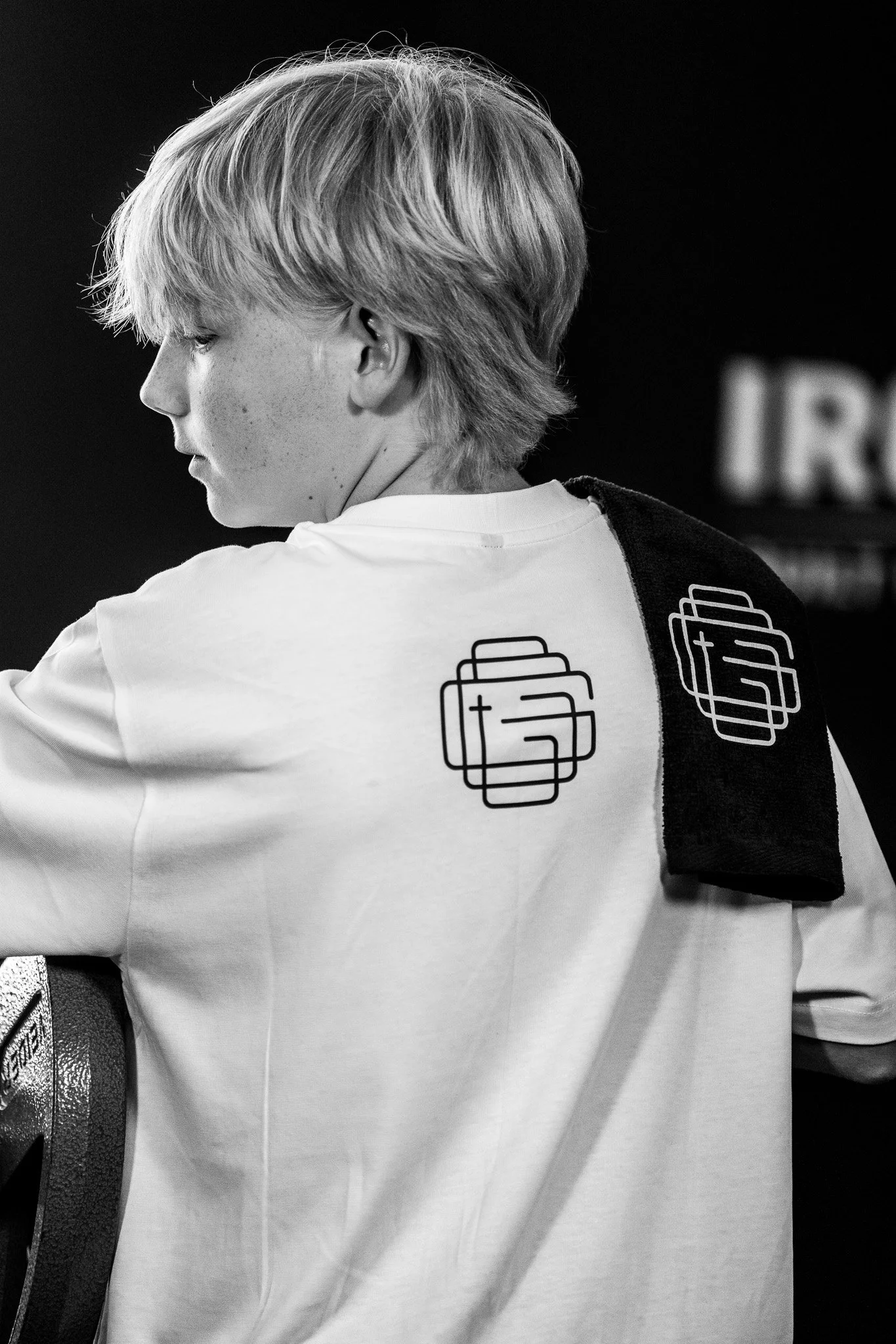A black and white photo of a young boy with light-colored hair in profile, wearing a white shirt with a logo on the back, with a black gym towel over his shoulder.
