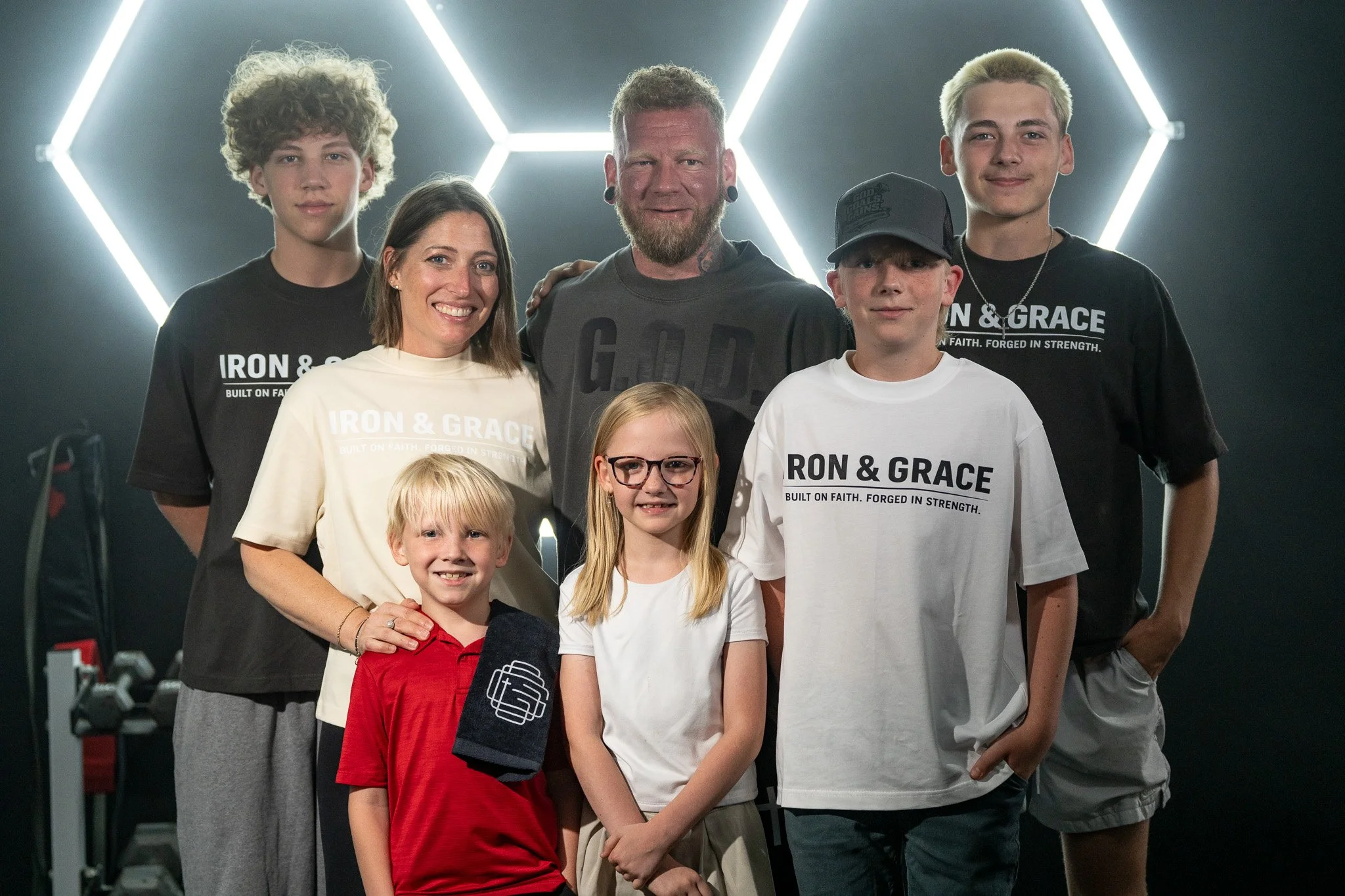 Group photo of seven people, including parents and children, standing together in a gym with neon lights in the background. The individuals are smiling and looking at the camera, some wearing matching t-shirts with the words 'IRON & GRACE' and a logo.