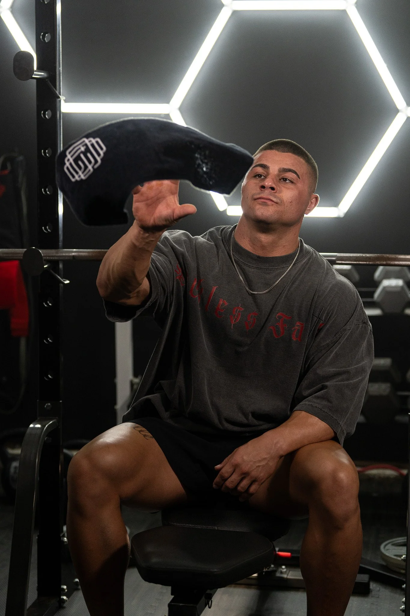 A man sitting on a chair in a gym, holding a black gym towel in his right hand, with a serious expression, wearing a dark gray T-shirt and shorts, surrounded by gym equipment and illuminated hexagonal ceiling lights.