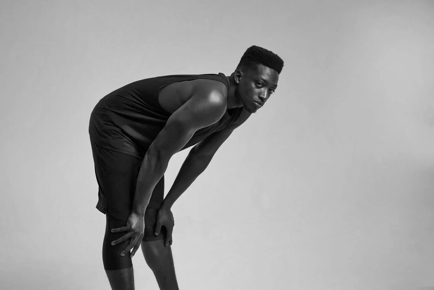 A young man in athletic clothing bends forward, with one hand resting on his thigh and the other on his knee, looking towards the camera against a plain background.