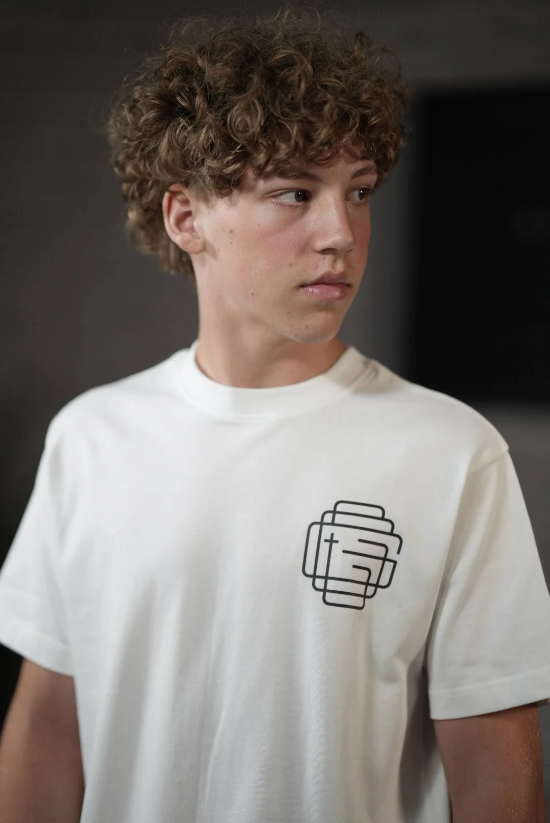 A young man with curly hair wearing a white T-shirt with a black geometric design on the chest, looking to the right. The background is dark and blurred.