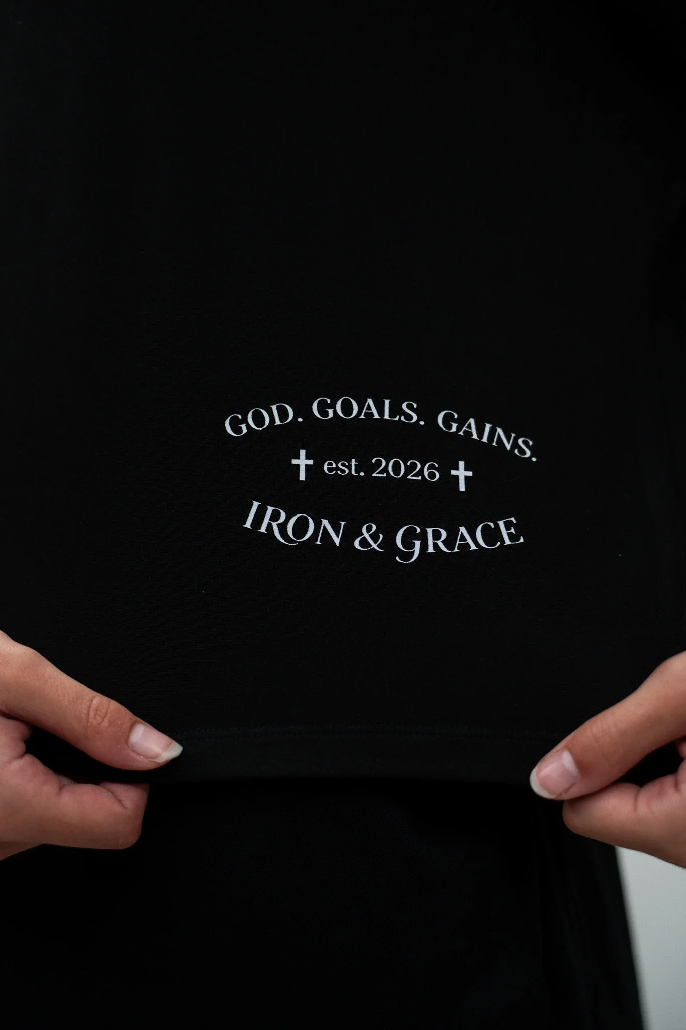 Close-up of a person's hands holding a black shirt with white text and symbols: 'GOD. GOALS. GAINS.', a cross symbols, 'est. 2026', and 'IRON & GRACE'.
