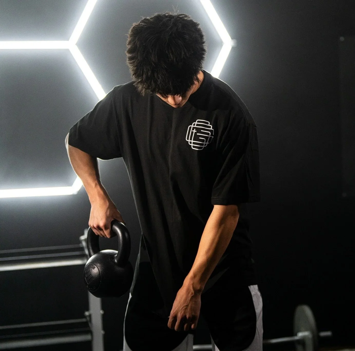 A young man with black hair lifting a kettlebell in a gym with geometric light fixtures on the wall.