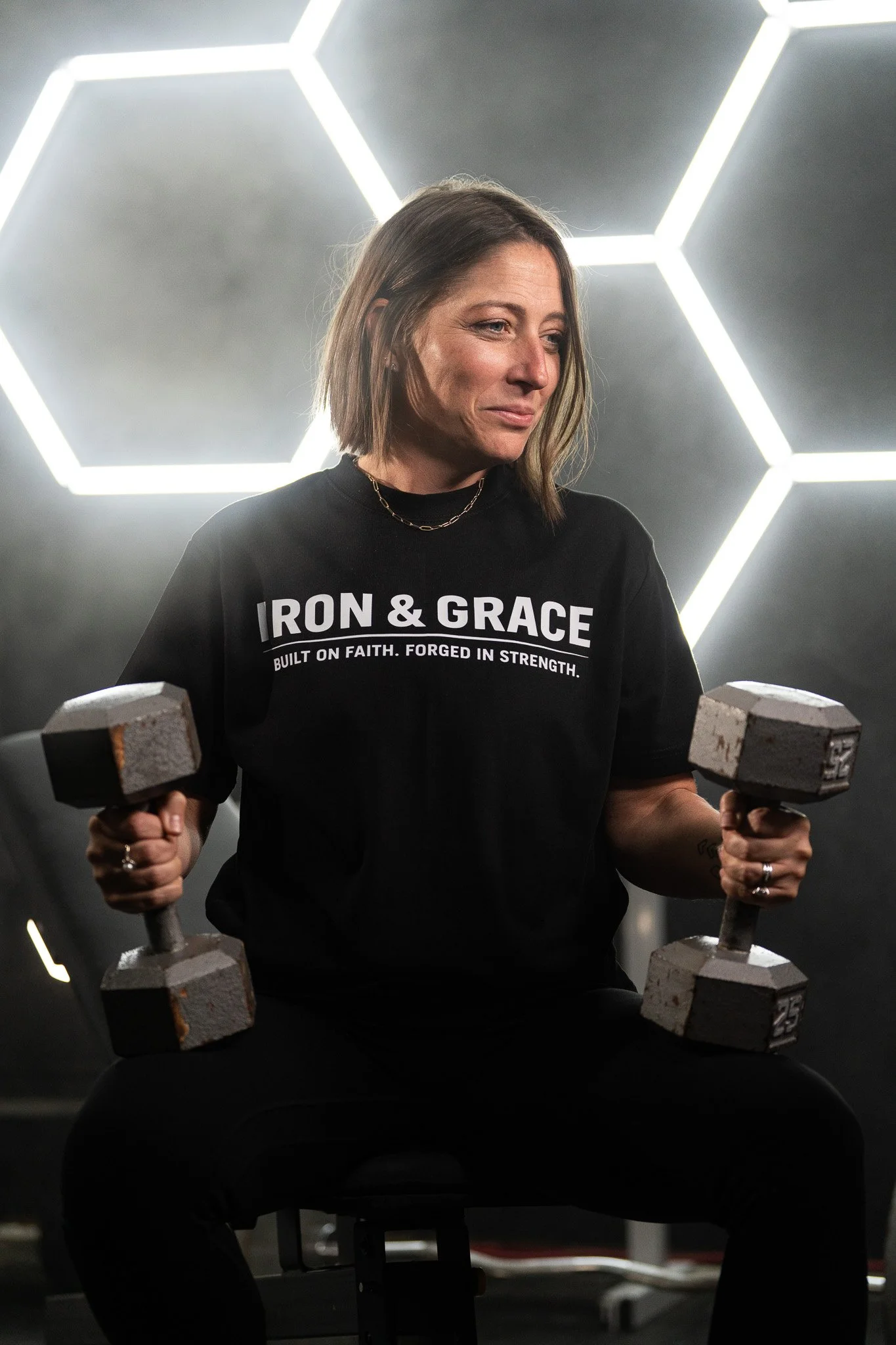Woman with shoulder-length hair lifting dumbbells in a gym, wearing a black T-shirt with the words 'IRON & GRACE BUILT ON FAITH. FORGED IN STRENGTH.' In a room with hexagon-shaped luminous ceiling lights.