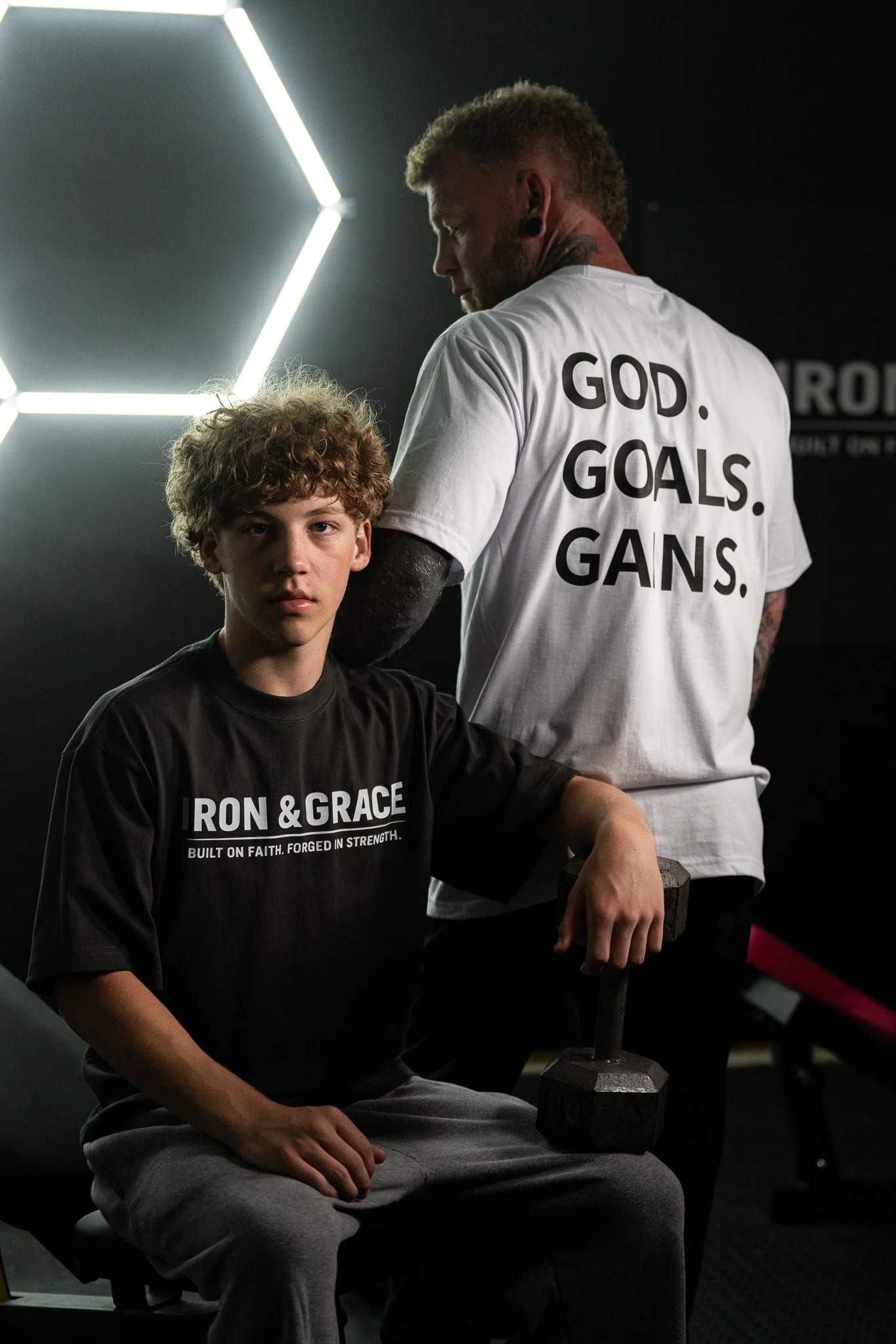 A young man with curly hair, wearing a black Iron & Grace t-shirt and gray sweatpants, sitting in a gym. Behind him, a man with a beard, wearing a white T-shirt with the words 'GOD. GOALS. GANS.' on the back, stands near a dumbbell, holding a piece of equipment in his left hand. The gym has black walls and modern lighting.