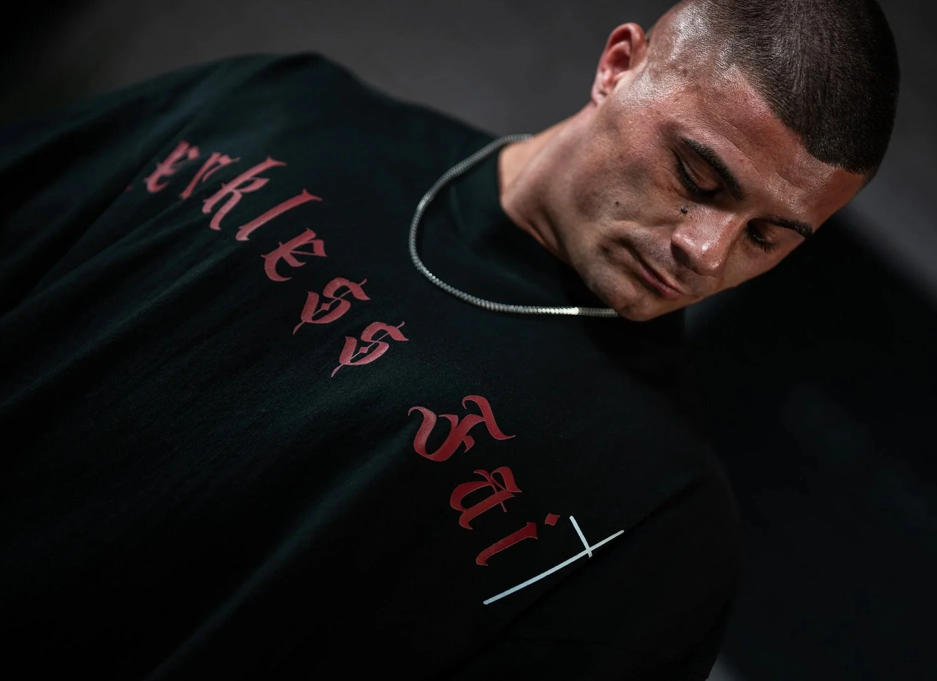 A young man with short hair and a chain necklace is looking down, wearing a dark T-shirt with red printed text and graphics, against a black background.