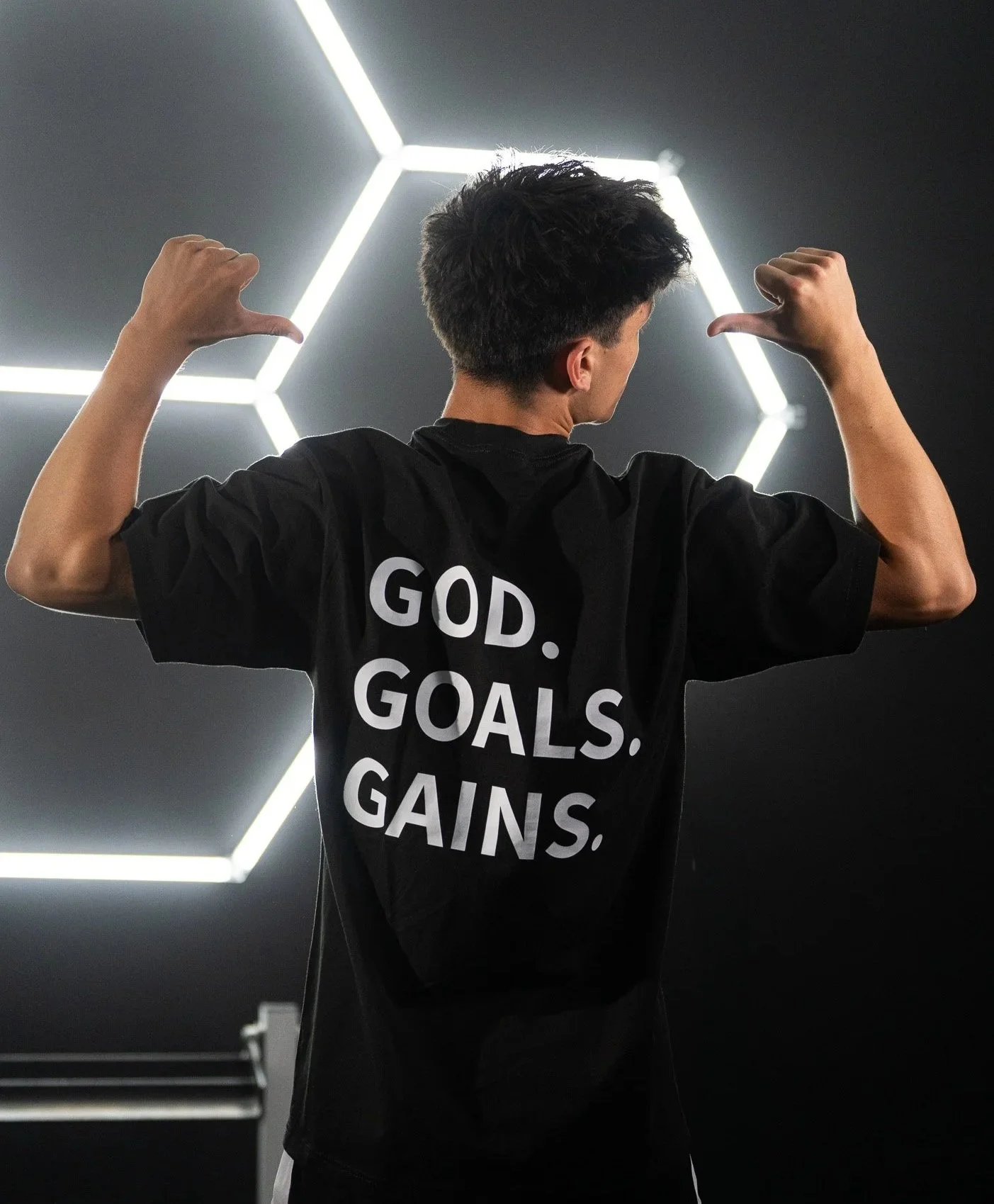 Young man flexing his biceps in front of a modern geometric light installation, wearing a black T-shirt that says 'GOD. GOALS. GAINS.'