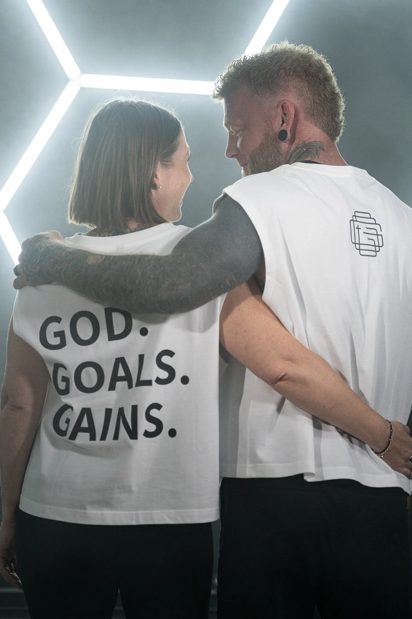 A man and a women are standing close together, smiling and touching shoulders in front of a modern ceiling light fixture. They are wearing white shirts with motivational phrases and black pants, in a bright, indoor setting.