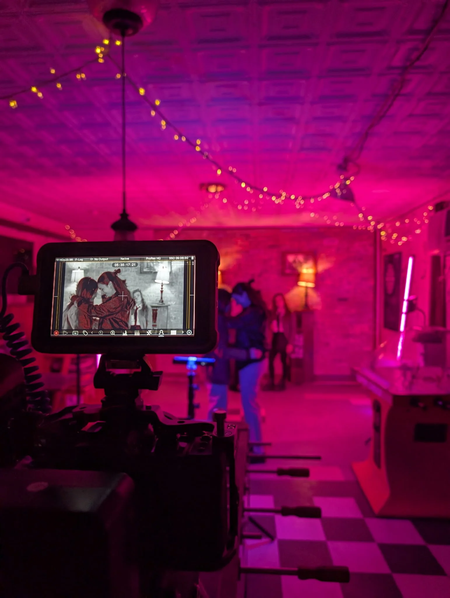 The image shows a room with purple lighting and string lights hanging from the ceiling. People are gathered in the background, and there is a camera on a tripod in the foreground, recording or taking photos. The floor has a checkered pattern and there are some chairs and furniture in the room.
