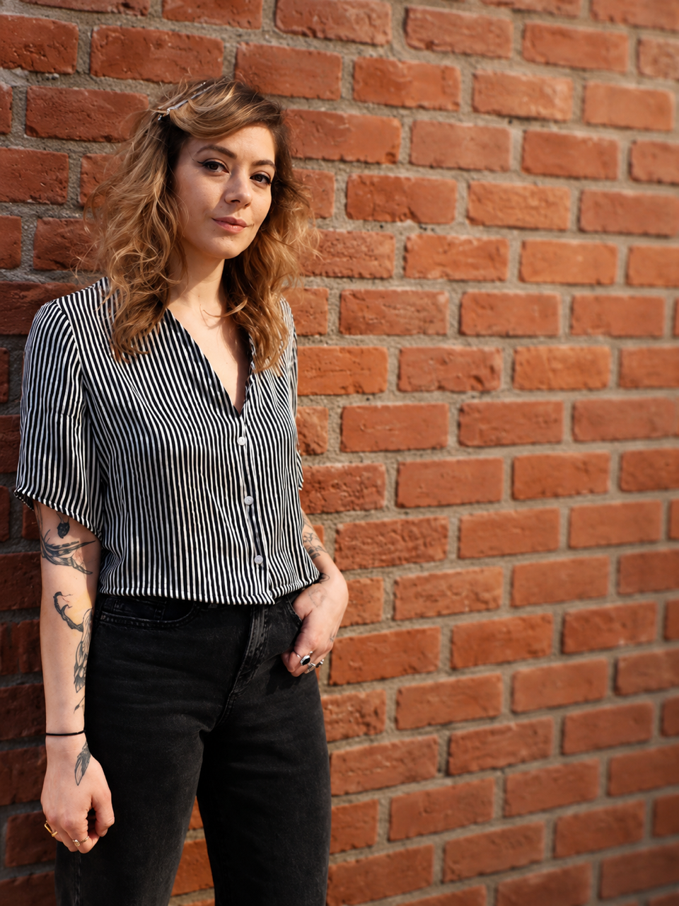 A young woman with wavy, shoulder-length blonde hair and tattoos on her left arm, wearing a black and white striped blouse and black jeans, standing against a red brick wall.