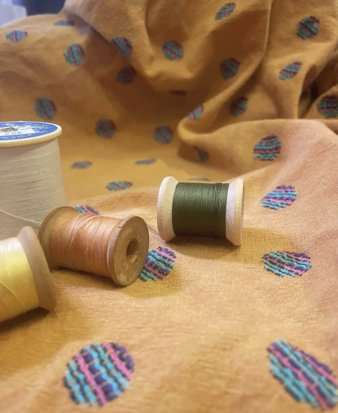 Three wooden spools with yellow, pink, and green thread, placed on yellow fabric with colorful polka dot pattern.