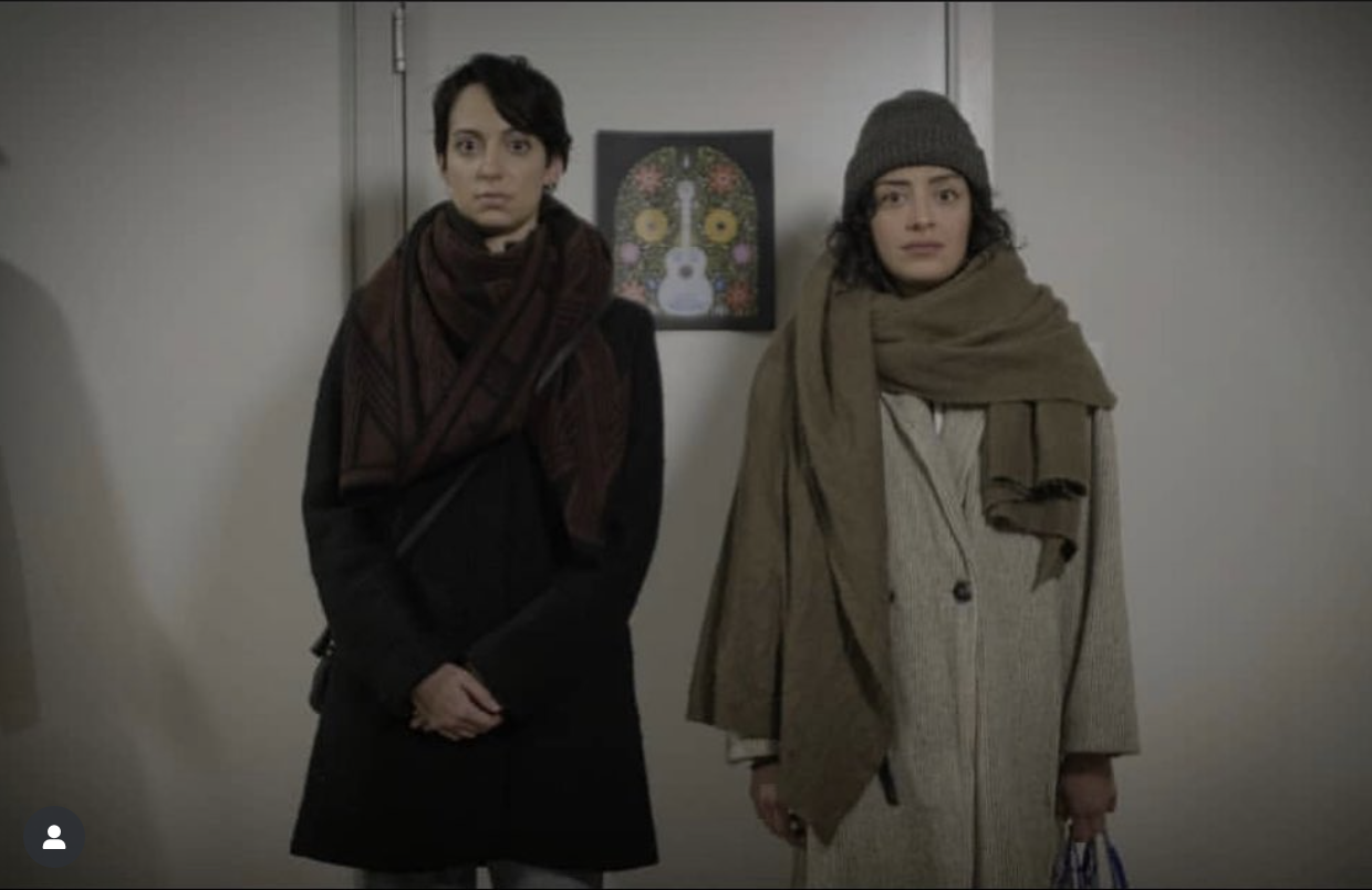 Two women standing against a wall with a colorful musical instrument and floral artwork in the background. Both women are dressed in winter coats and scarves.