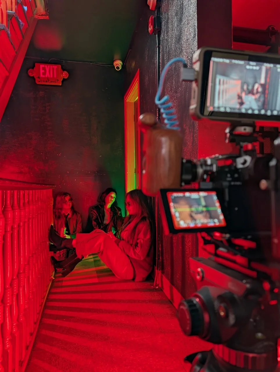 Three women sitting on a staircase in a dimly lit space with red lighting, a camera on a tripod capturing the scene, and an exit sign in the background.