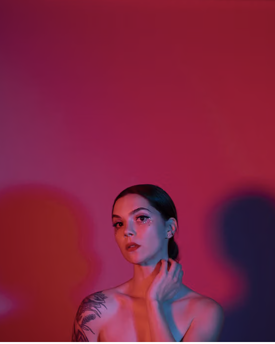 Portrait of a woman with dark hair, makeup with rhinestones under her eye, and a visible tattoo on her shoulder, set against a colorful blurred background.