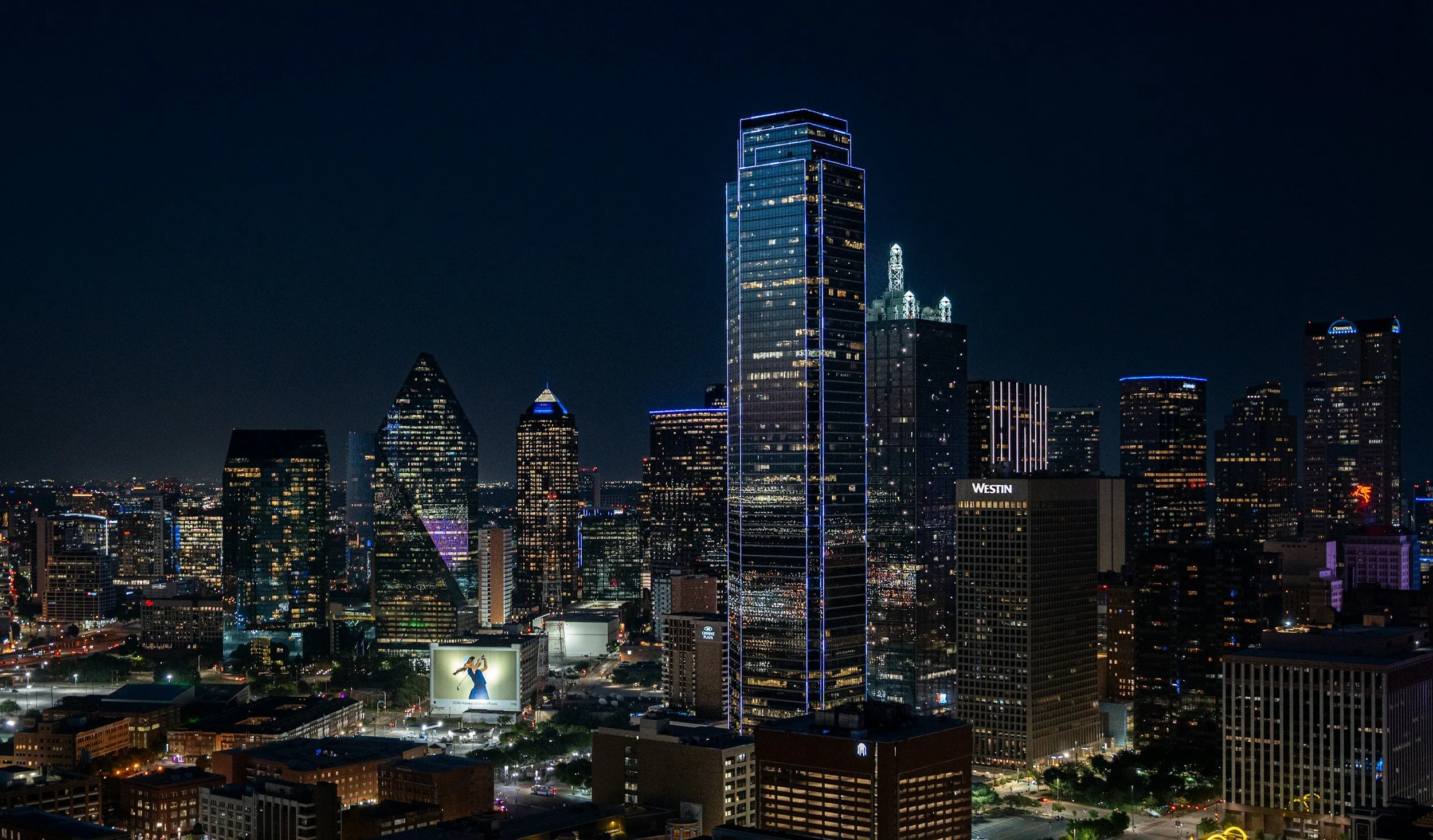 Nighttime cityscape with high-rise buildings and illuminated windows, including a tall building with blue light accents and the Westin hotel sign, and city lights below.