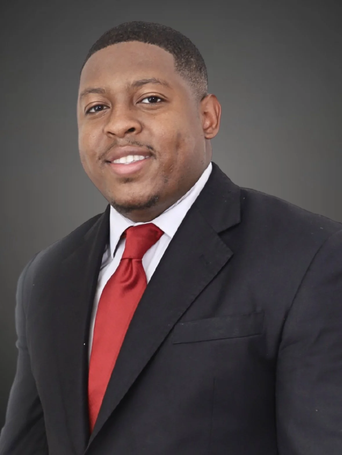 A man wearing a black suit, white shirt, and red tie posing against a gray background.