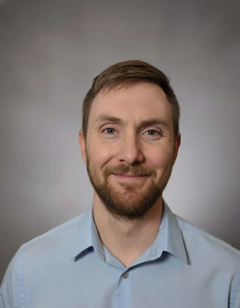 A man with short brown hair and a beard, smiling, wearing a light blue collared shirt against a gray background.