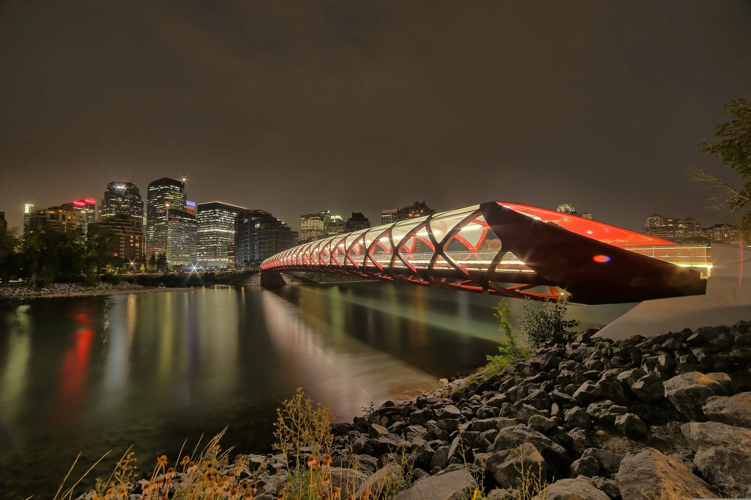 Calgary's illuminated Peace Bridge spanning the Bow River at night, with the downtown skyline in the background.
