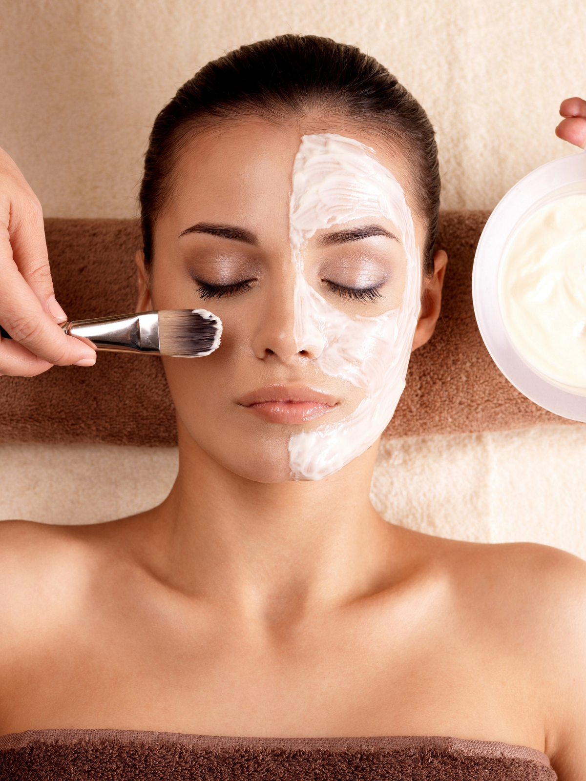 A woman receiving a facial treatment, with half her face covered in a white cream mask, while a technician applies cream with a brush. The woman is lying down with closed eyes.