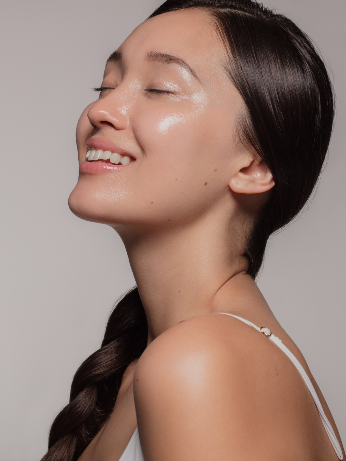 A woman with smooth skin and long, dark hair styled in a braid, smiling with closed eyes and showing her teeth, against a plain background.