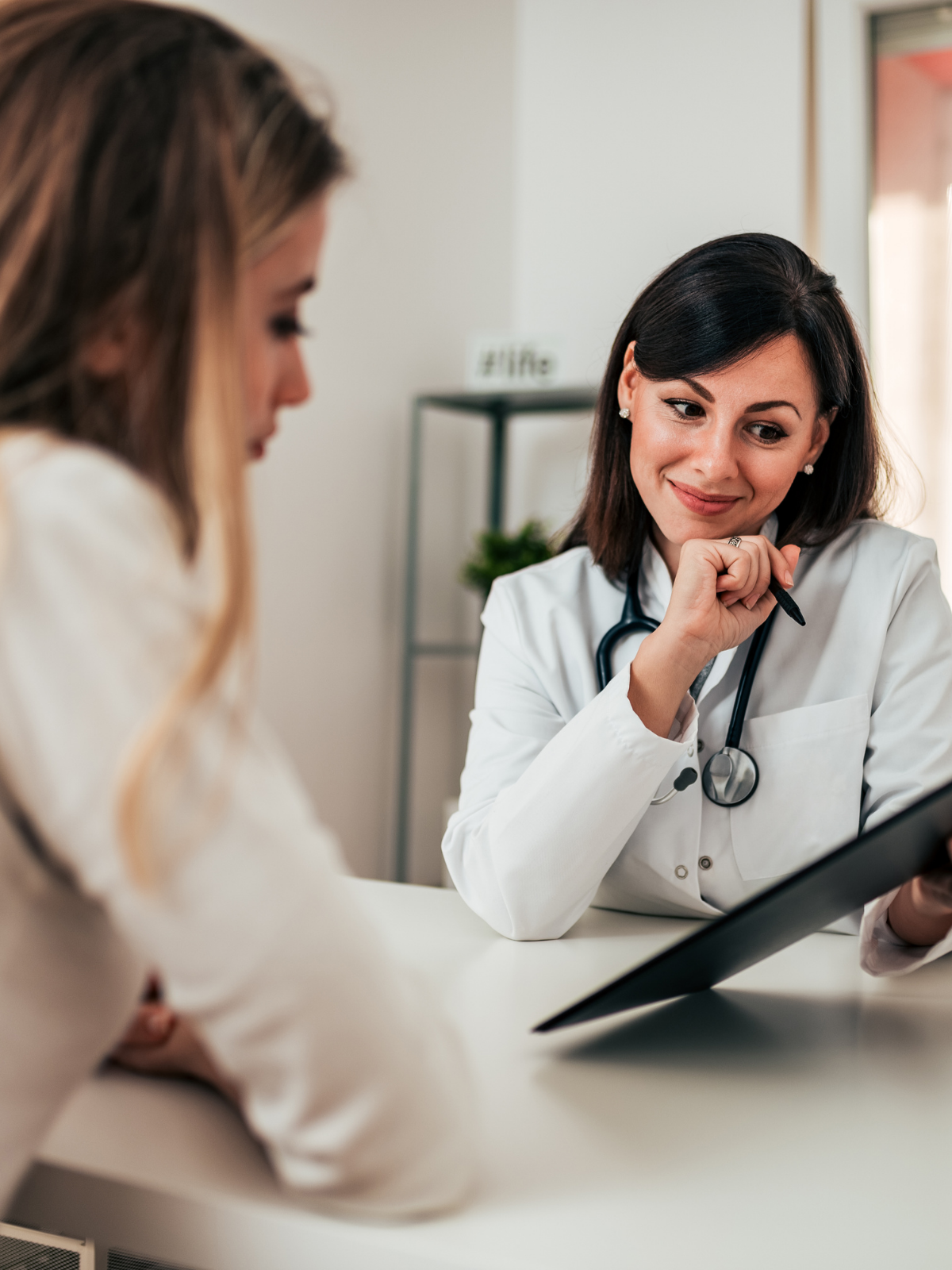 A female doctor with dark hair and a stethoscope around her neck smiling and looking at a tablet, sitting with a young woman with blonde hair in a medical office.
